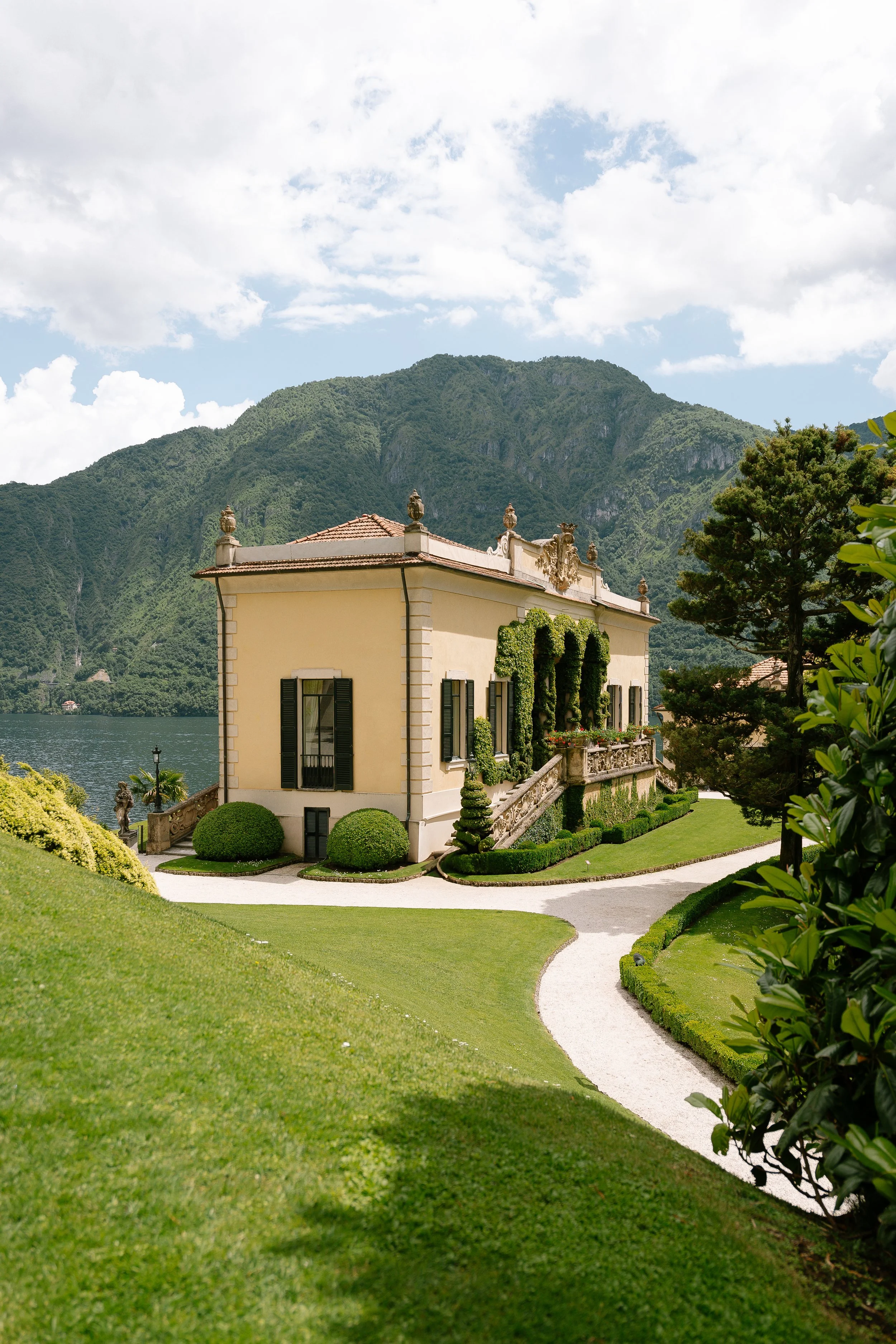 A large, yellow mansion with shutters on the windows, surrounded by a well-maintained garden with curved pathways, greenery, and mountains in the background.