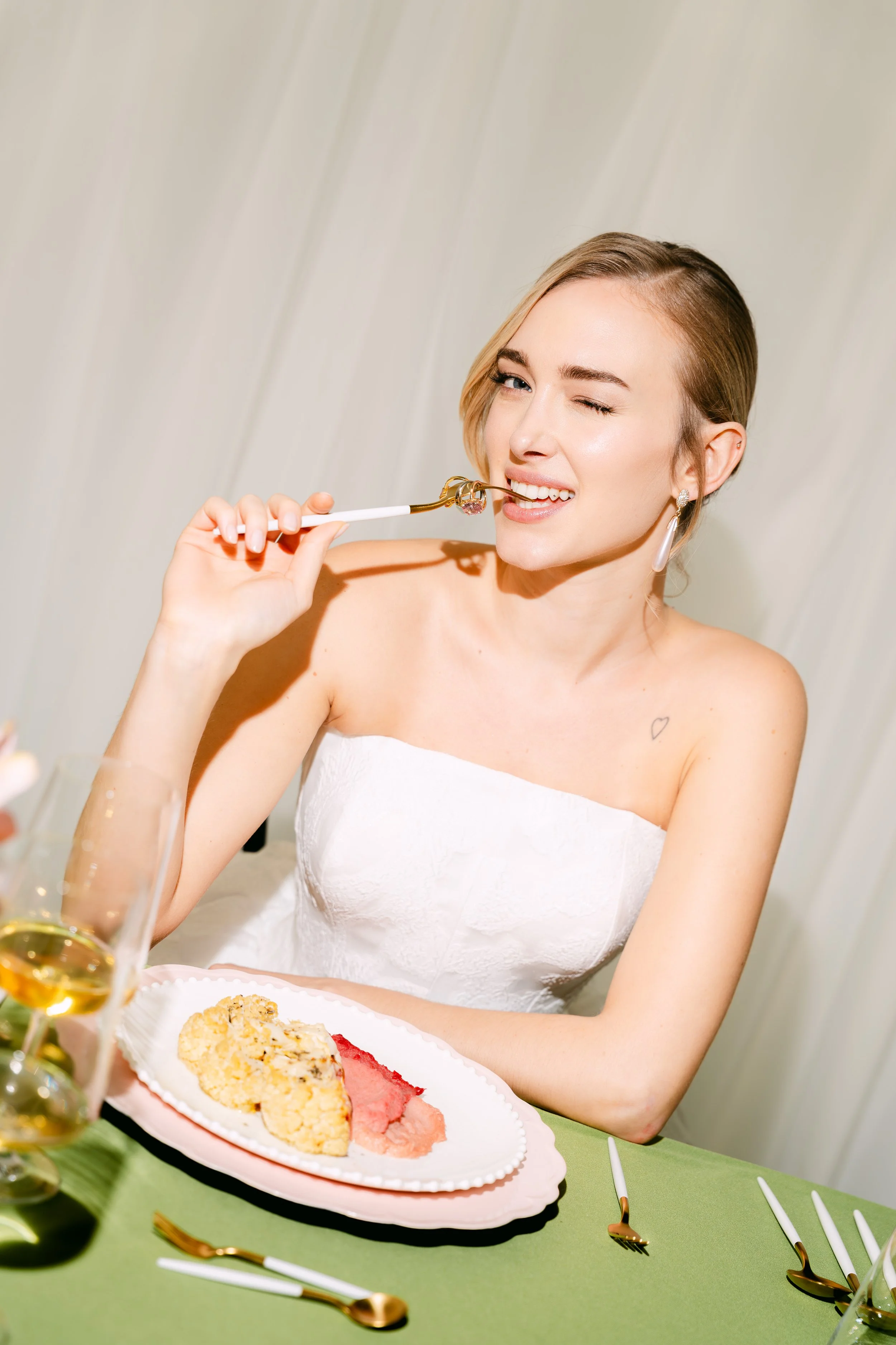 A woman with short blonde hair wearing a white strapless dress, holding a fork near her mouth, with a playful winking expression. She is sitting at a table with a green tablecloth, and there is a plate of dessert and a glass of white wine in the fore