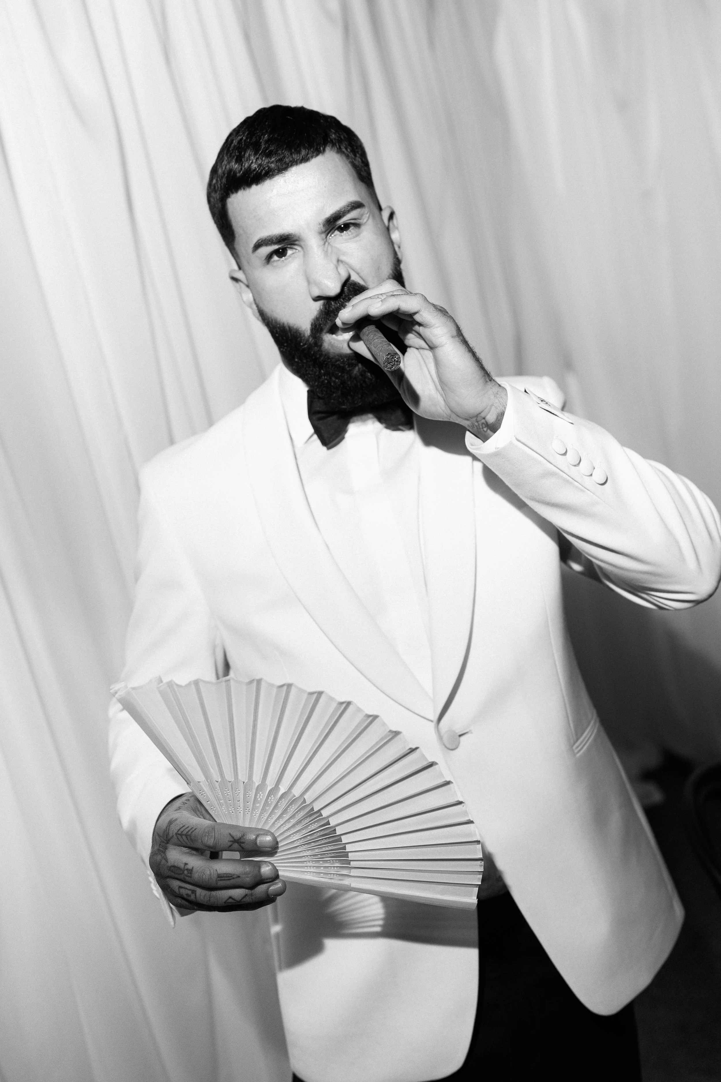 A man with a beard and tattoos, dressed in a white tuxedo with a black bow tie, holding a fan and a cigar, looking directly at the camera in front of a plain background.
