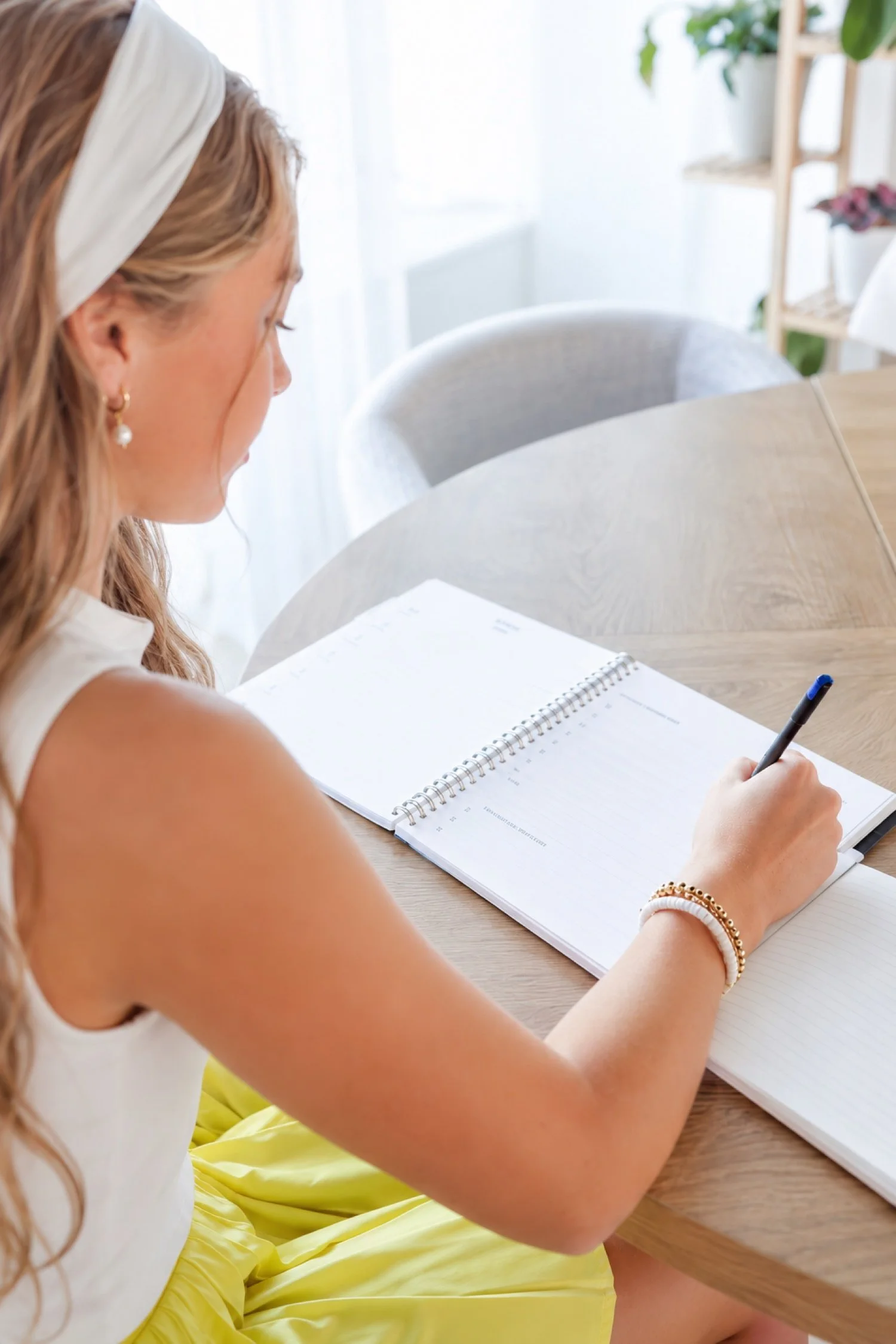A woman writes in a notebook at a table beside a window.