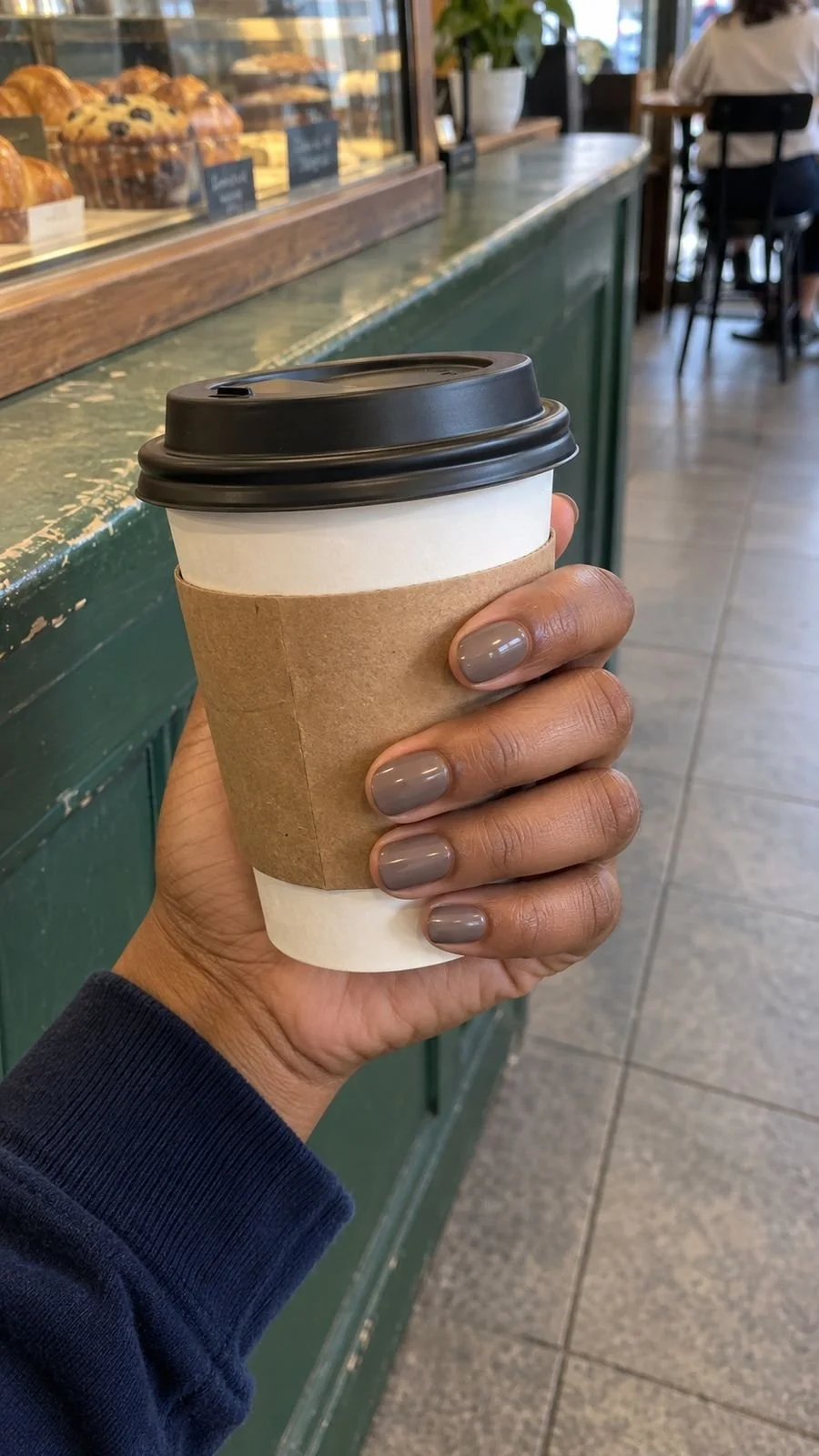 Taupe nails on a hand holding a closed coffee cup inside a cafe.