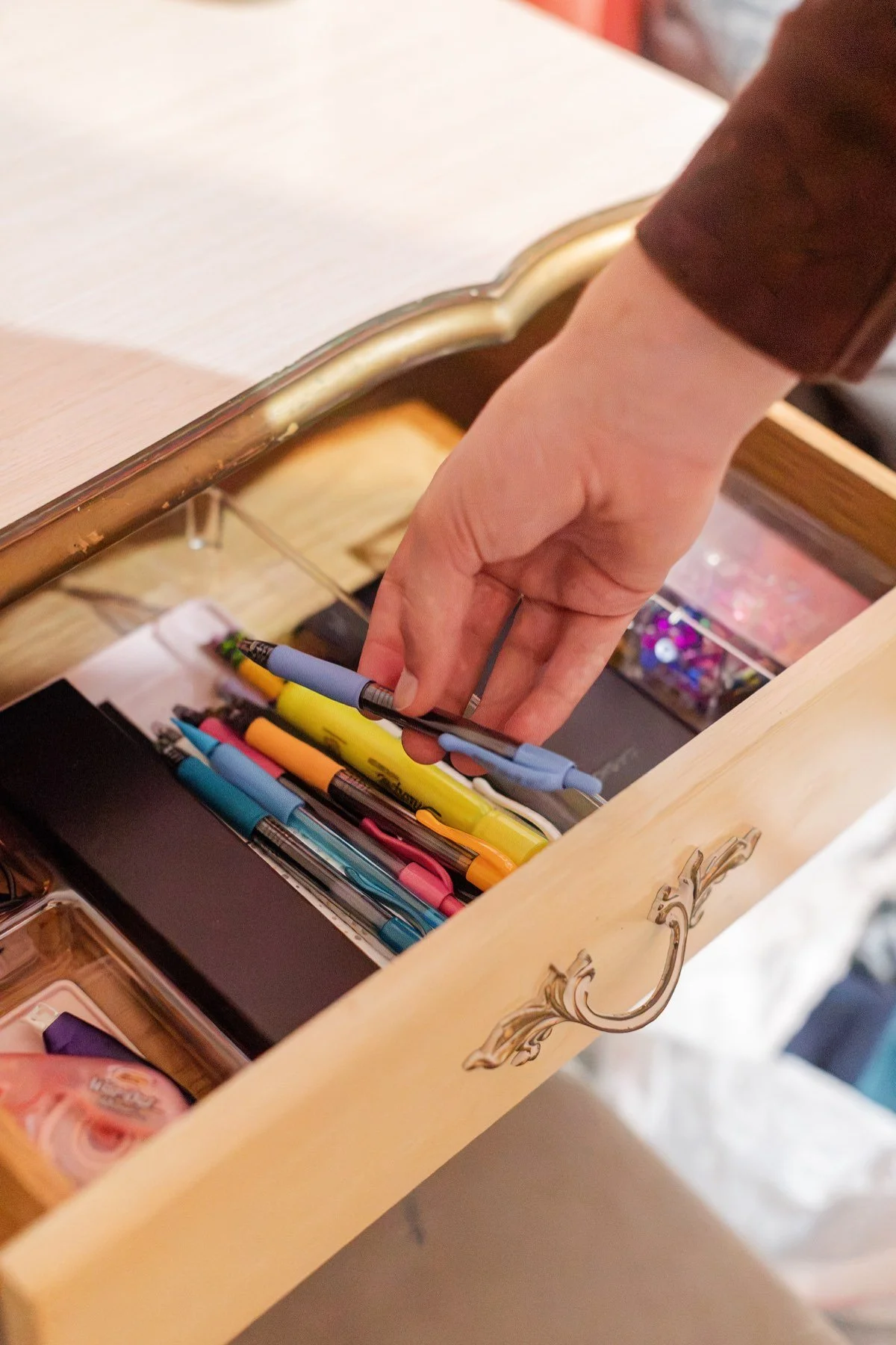 A hand picks up a blue pen from a clear tray inside a wooden desk drawer.