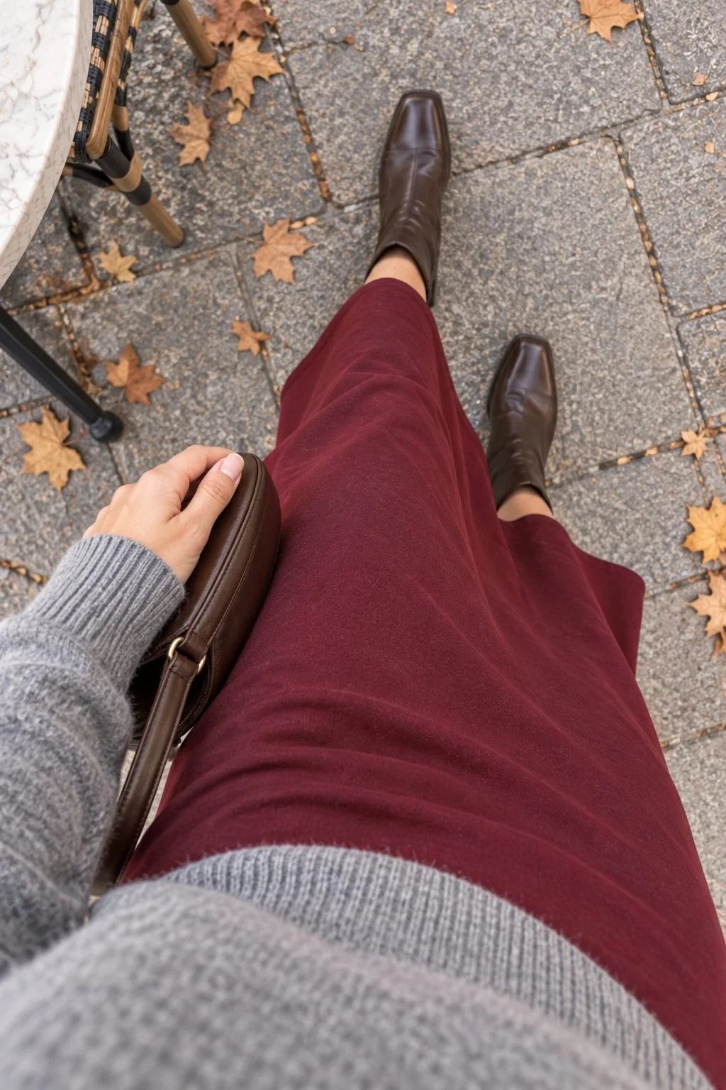 A wearer looks down at a burgundy midi skirt, brown boots, a gray sweater cuff, and a small brown bag.