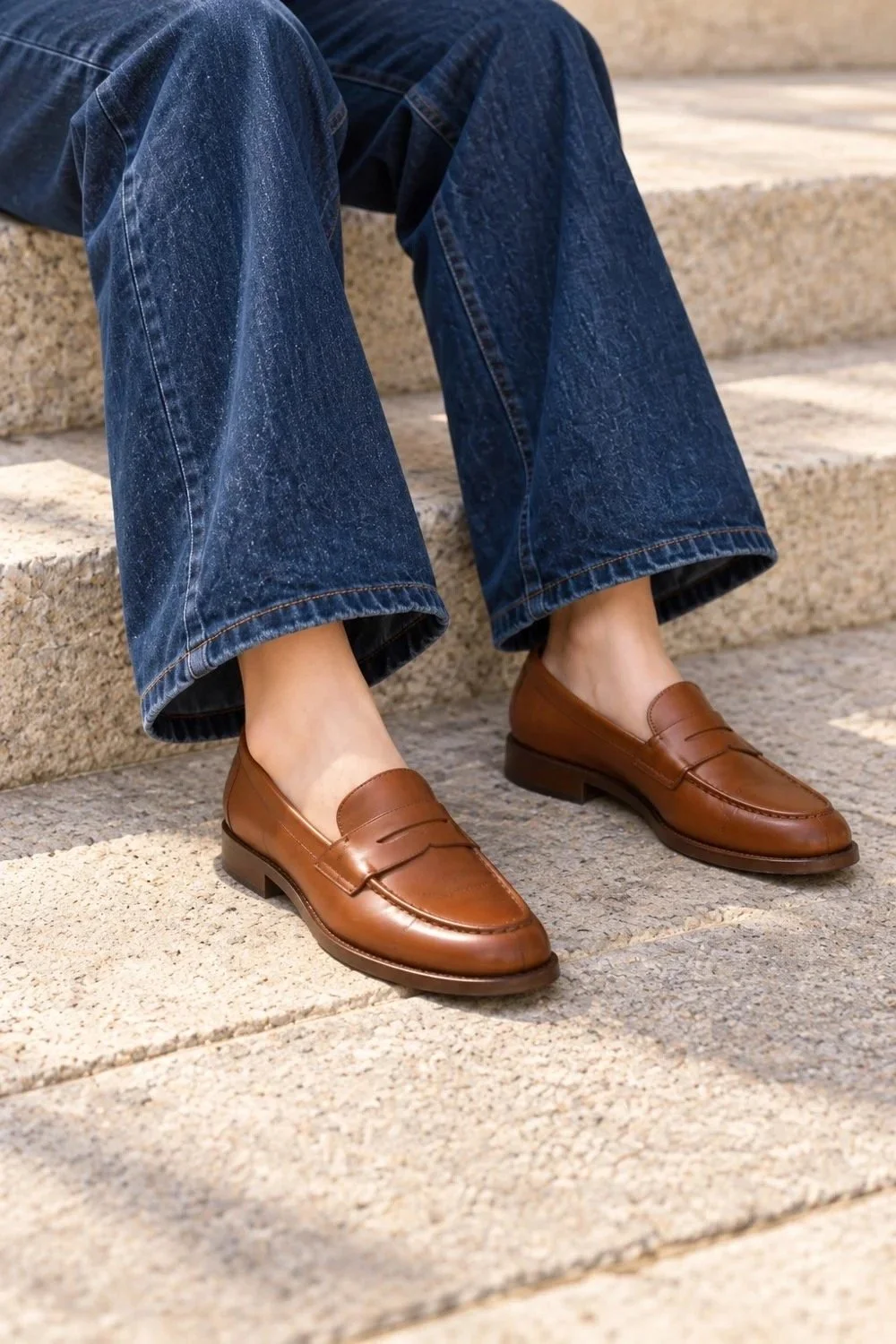 Close view of wide-leg dark denim hems and brown penny loafers on stone steps.