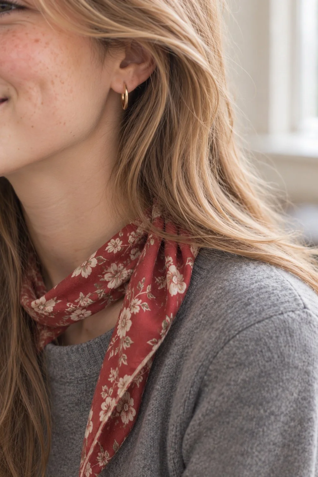 Gold hoop earring beside blonde hair, with a red scarf and gray sweater.