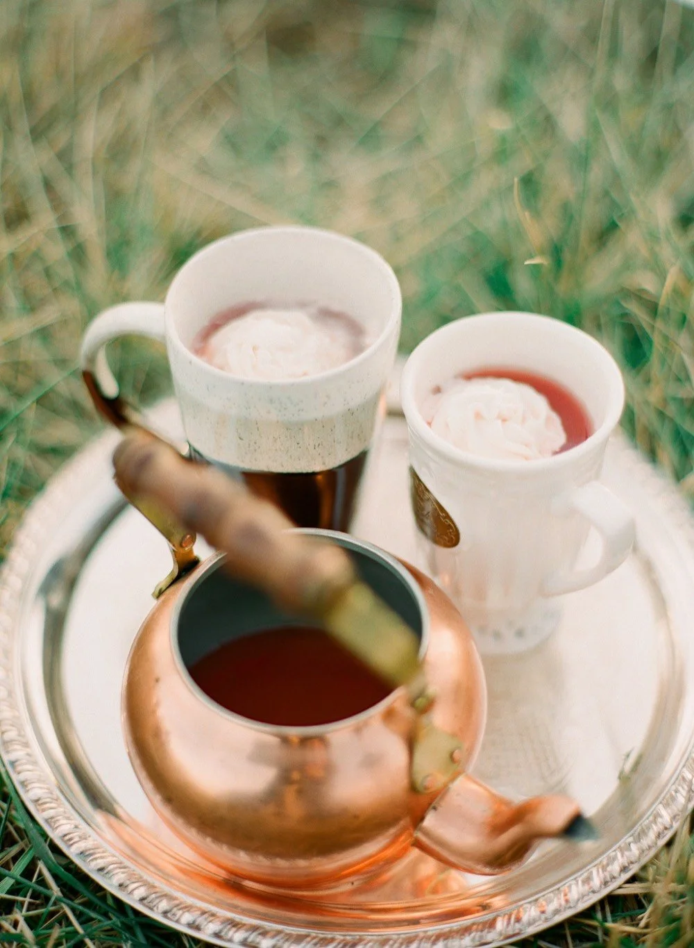 Two mugs of reddish wassail topped with whipped cream beside a copper-colored pot.
