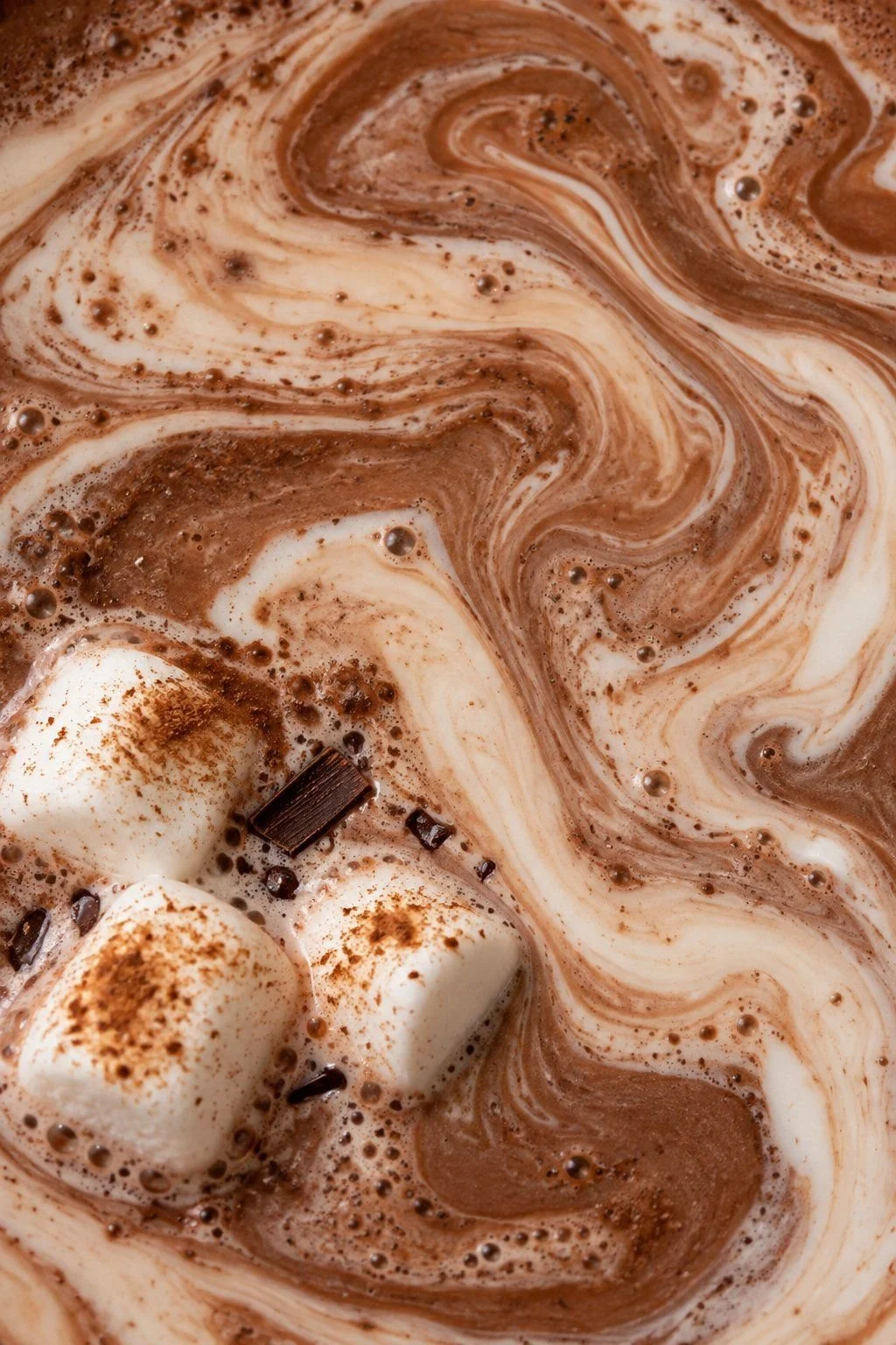Three cinnamon-dusted marshmallows beside pale cream waves in milk chocolate cocoa.