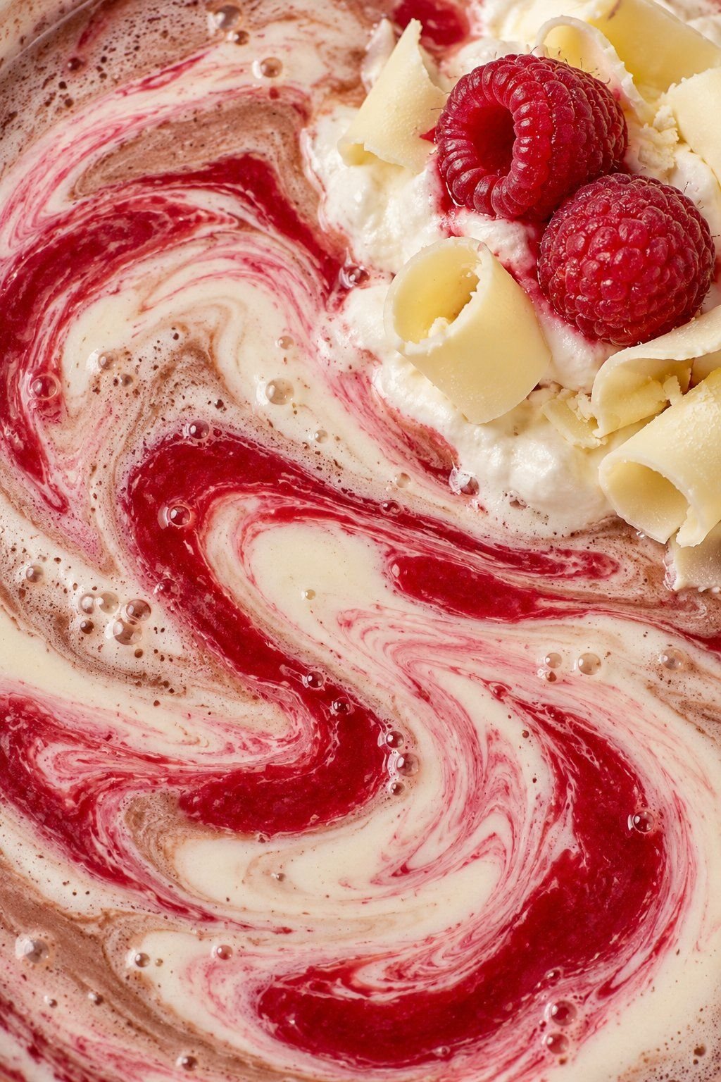 Two raspberries and white chocolate curls on cream beside red and ivory cocoa swirls.