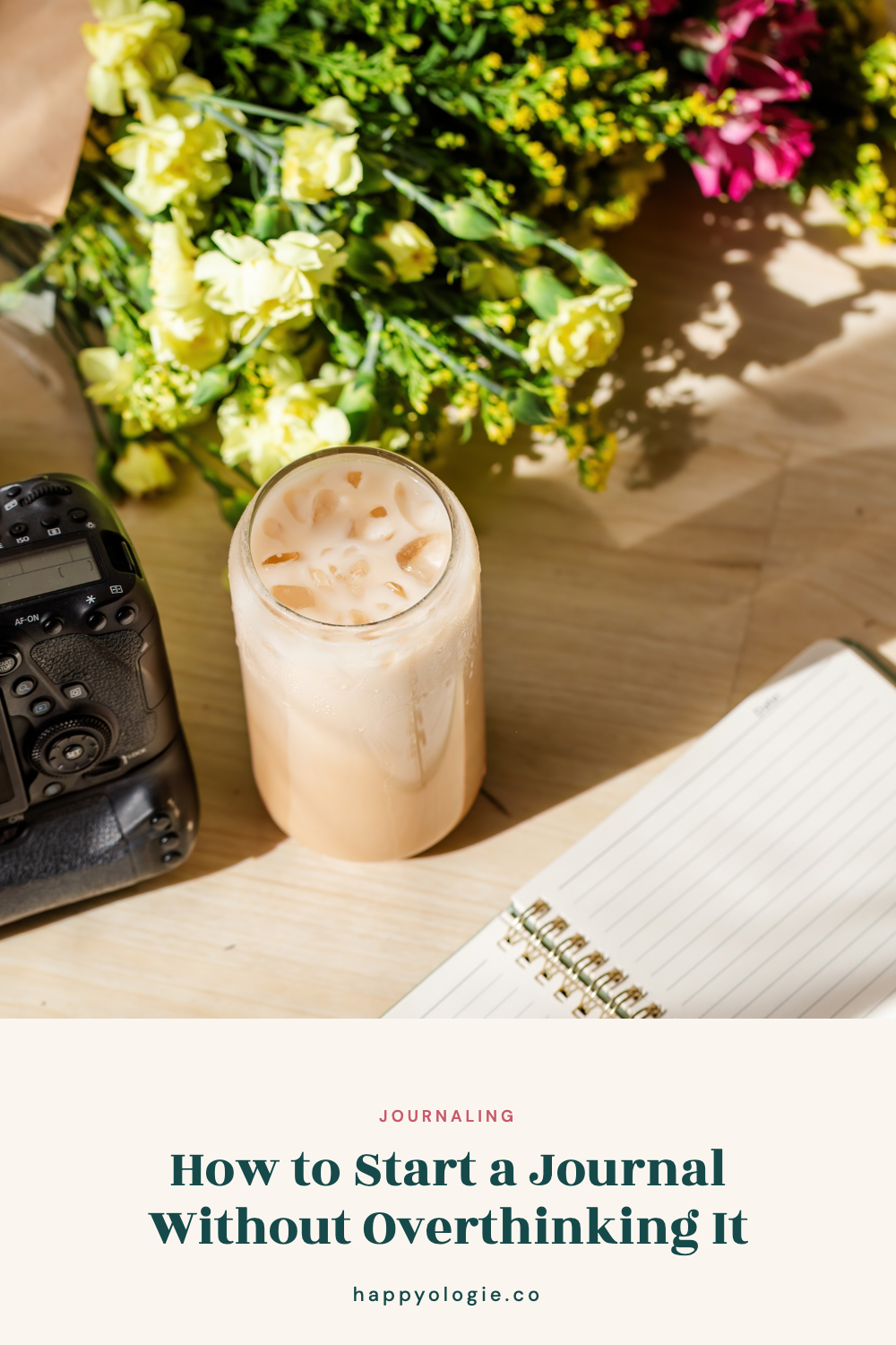 Open lined journal beside an iced drink, flowers, and a camera on a sunny desk.