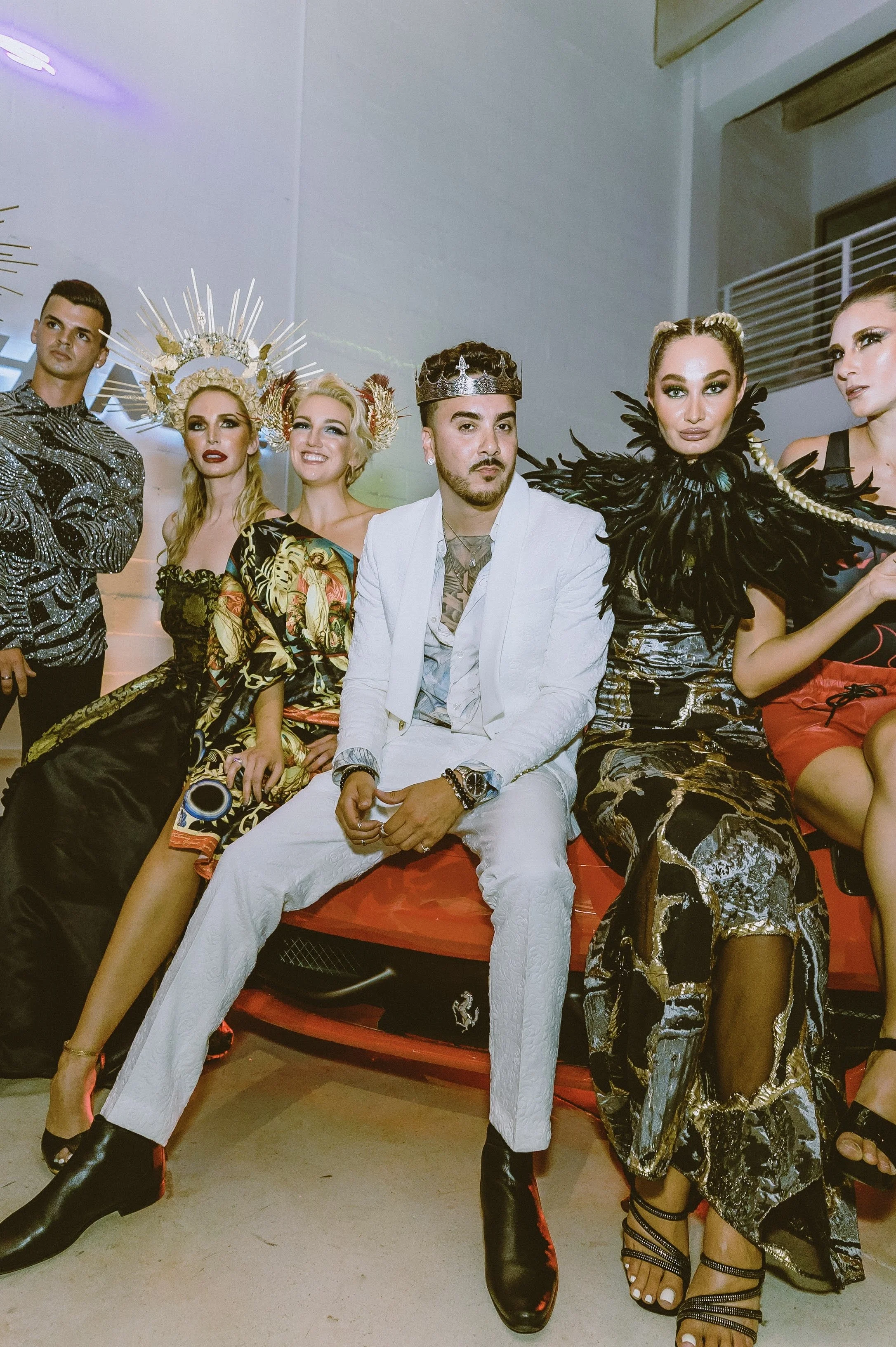A group of six fashionably dressed individuals sitting on a red couch at an event or fashion show, with some wearing bold makeup and jewelry, and two women wearing elaborate headpieces.