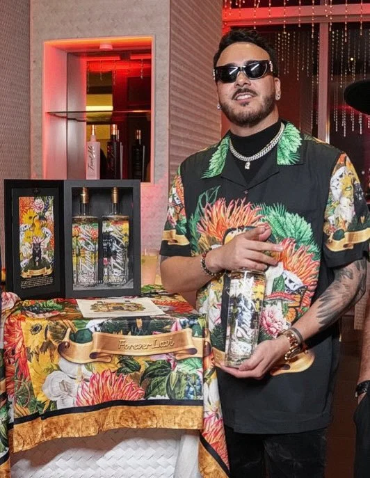Man wearing sunglasses and a colorful floral shirt holding a bottle, standing in front of a display with similar bottles and floral wrapping, in a decorated indoor setting with dim lighting.
