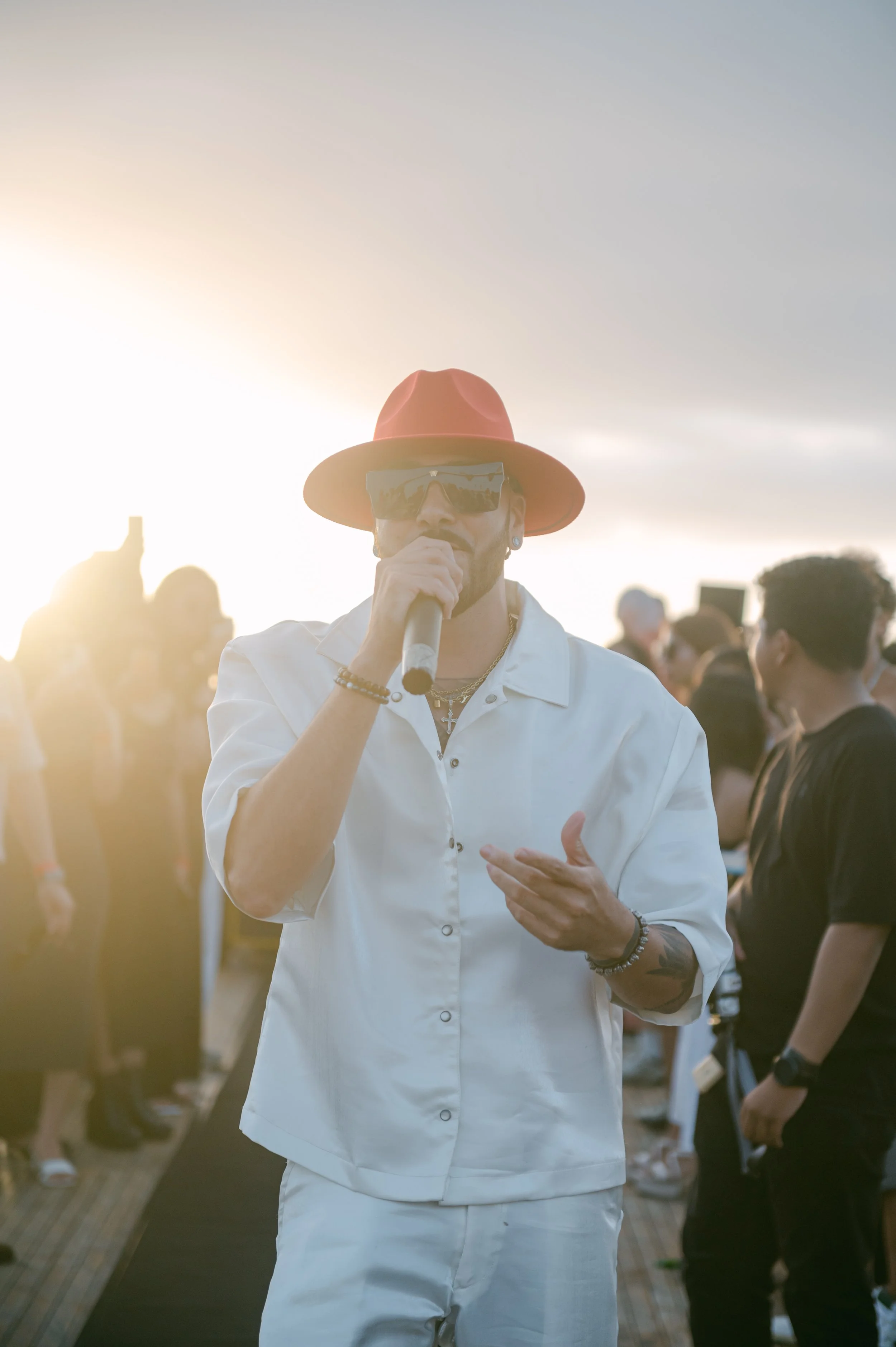 A man wearing a white shirt and white pants, red hat, and sunglasses, holding a microphone and singing at an outdoor event during sunset. There are people in the background.