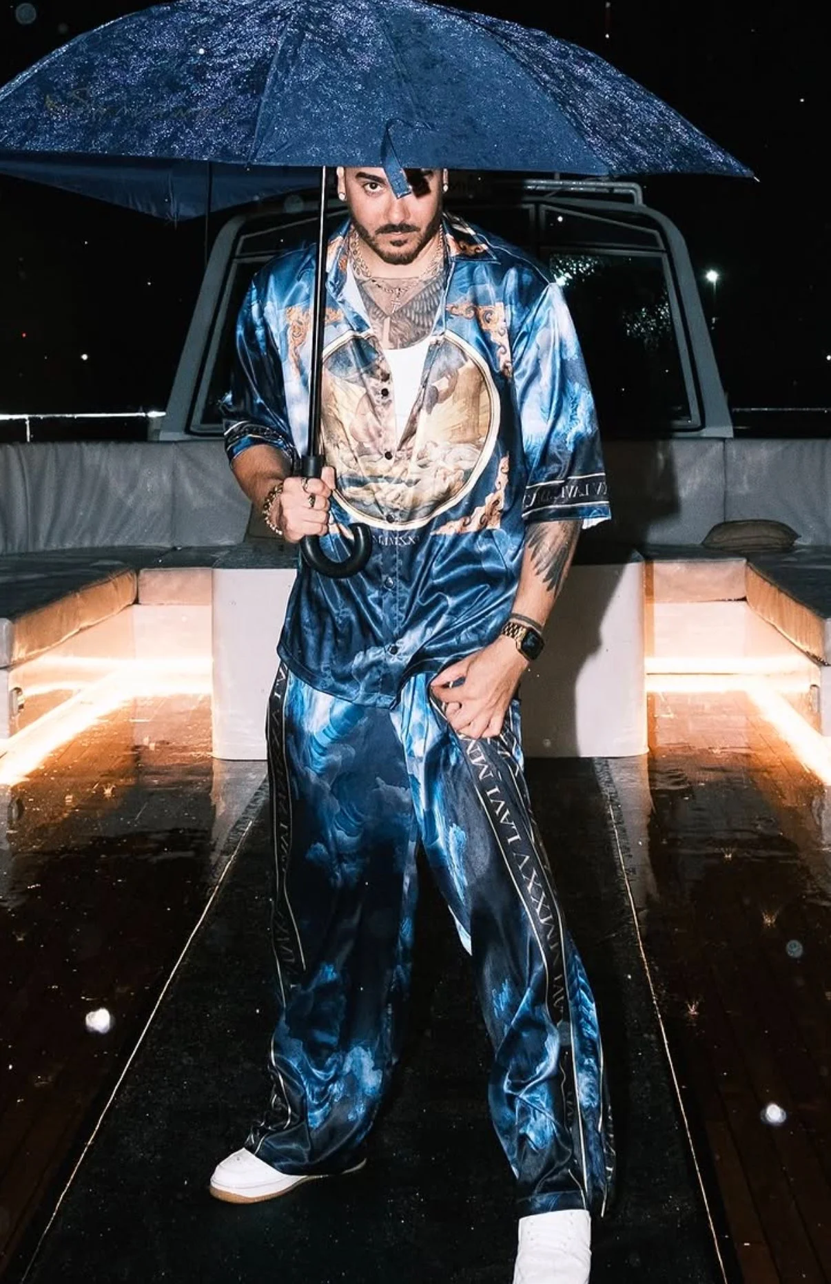 A man standing under a rain-covered umbrella in a stylish blue matching outfit with patterns, in a dimly lit outdoor setting at night.