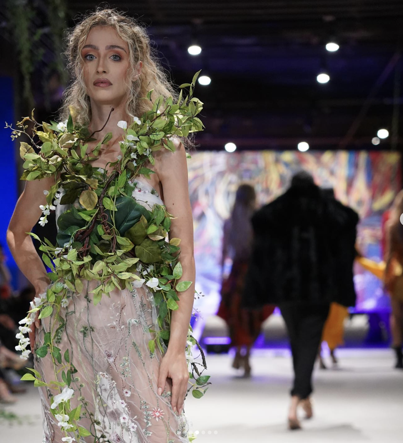 Model walking on runway wearing a dress with green leaves and white flowers, with a colorful abstract background and audience in the distance.