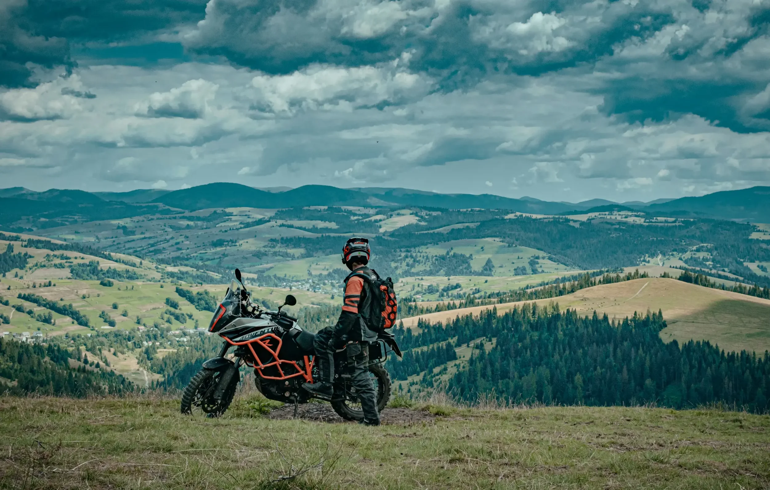 Person in motorcycle gear standing next to an adventure motorcycle on a grassy hilltop overlooking rolling green hills and a cloudy sky.