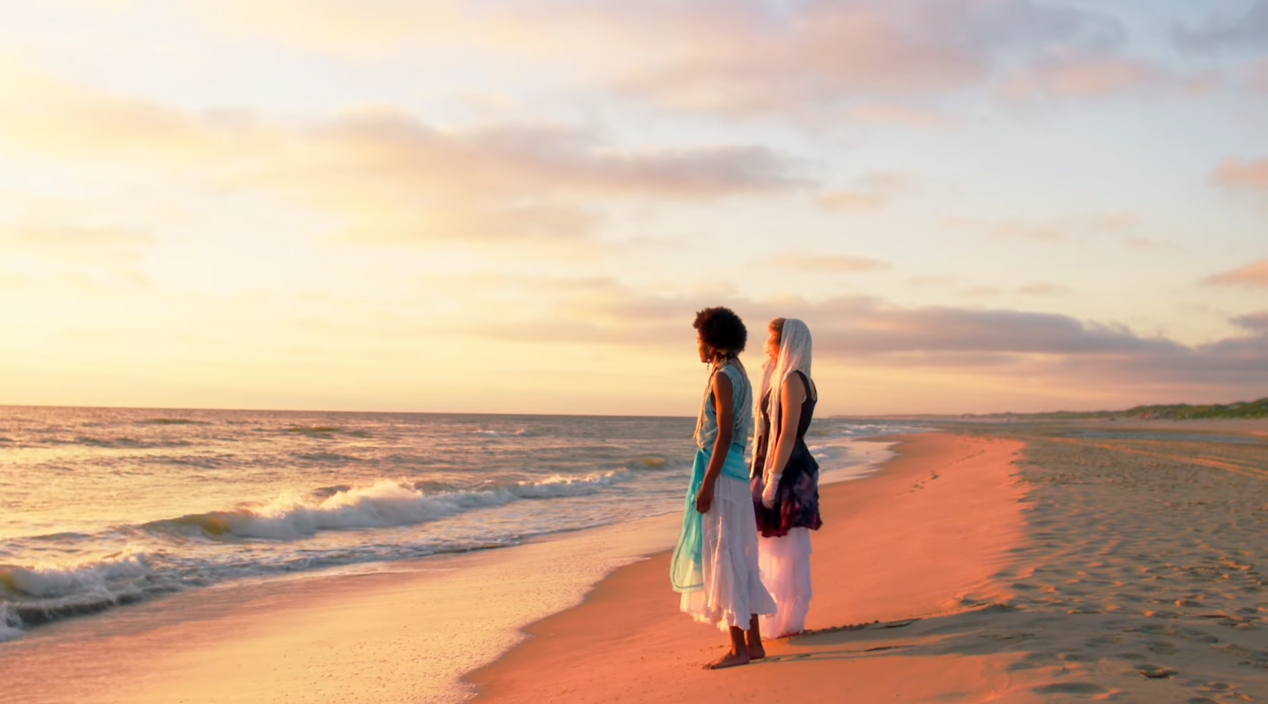Two women standing on a beach during sunset, facing the ocean, with light-colored skirts and head coverings, and the sky filled with clouds illuminated by the setting sun.
