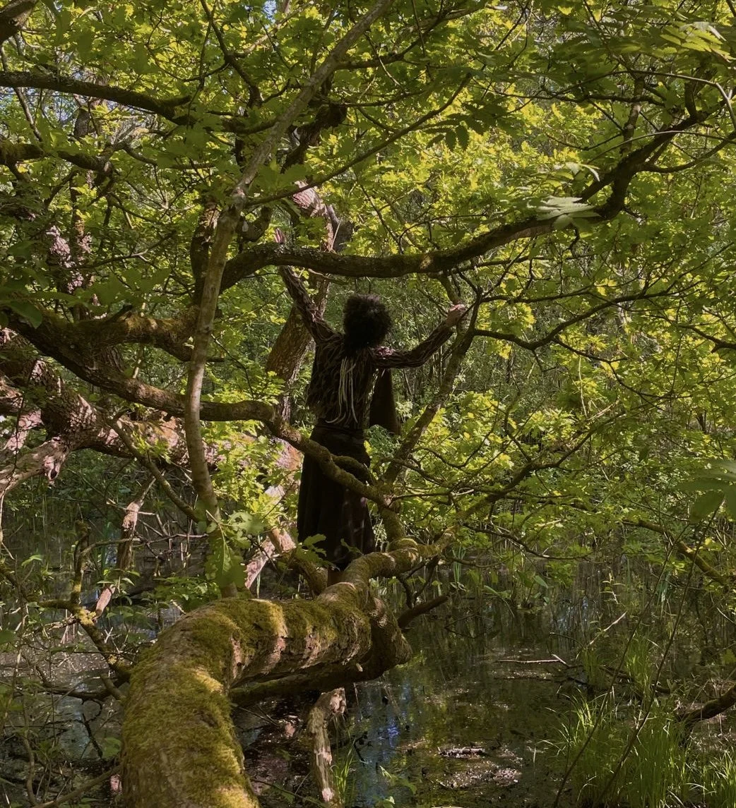 A person with curly hair wearing dark clothing standing on a moss-covered tree branch in a lush green forest, with their back facing the camera and surrounded by dense foliage.
