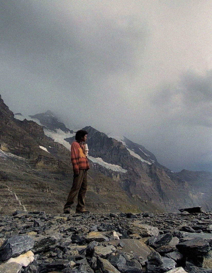 A person standing on rocky terrain with mountains and snow patches in the background under a cloudy sky.