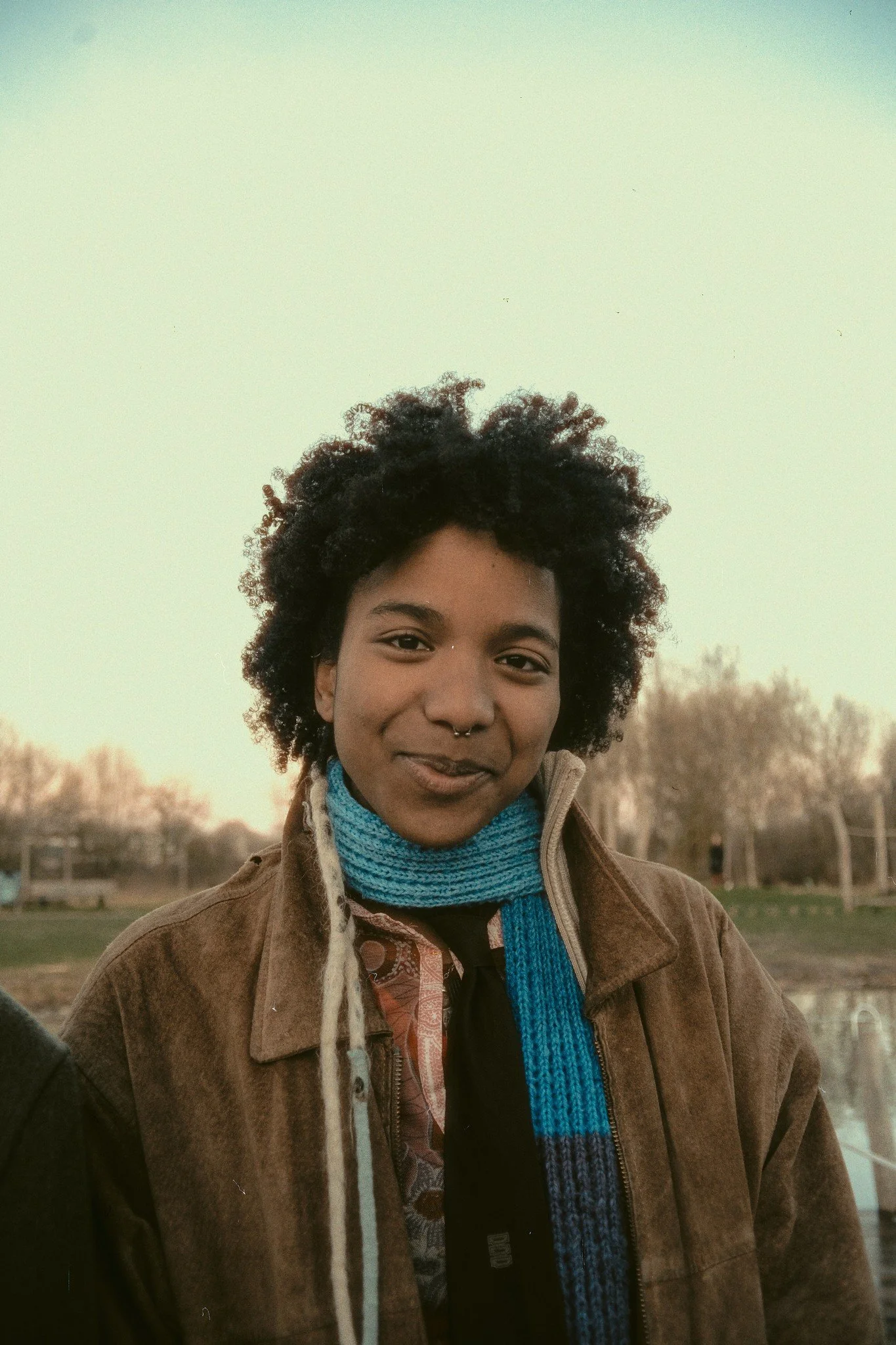 A person with curly hair, wearing a brown jacket, a blue knit scarf, standing outdoors near a body of water during sunset.