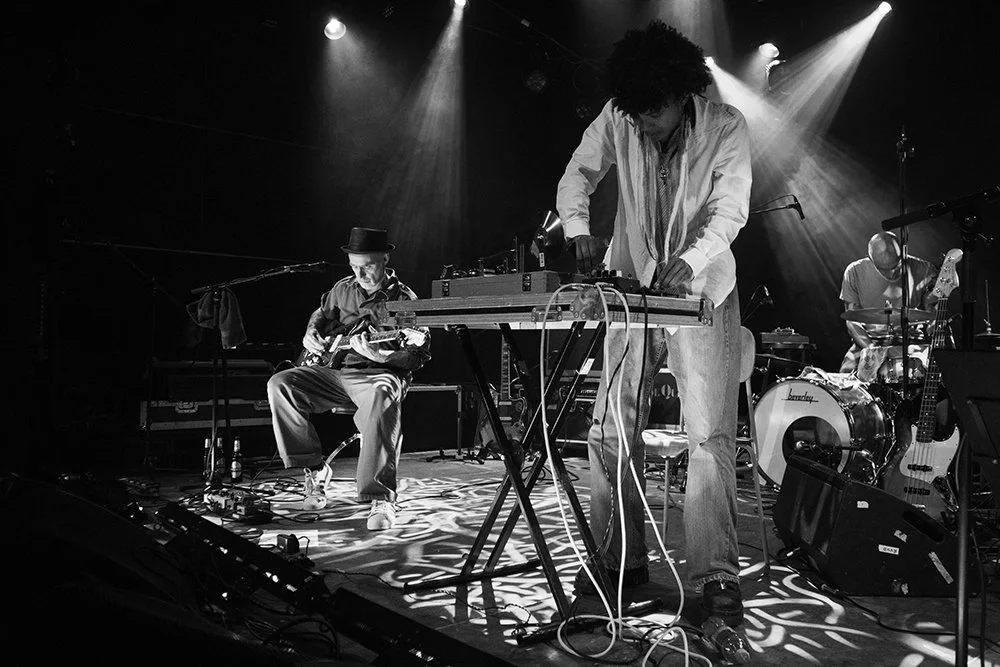 Four musicians performing on stage with various instruments, under stage lights, in a black and white image.
