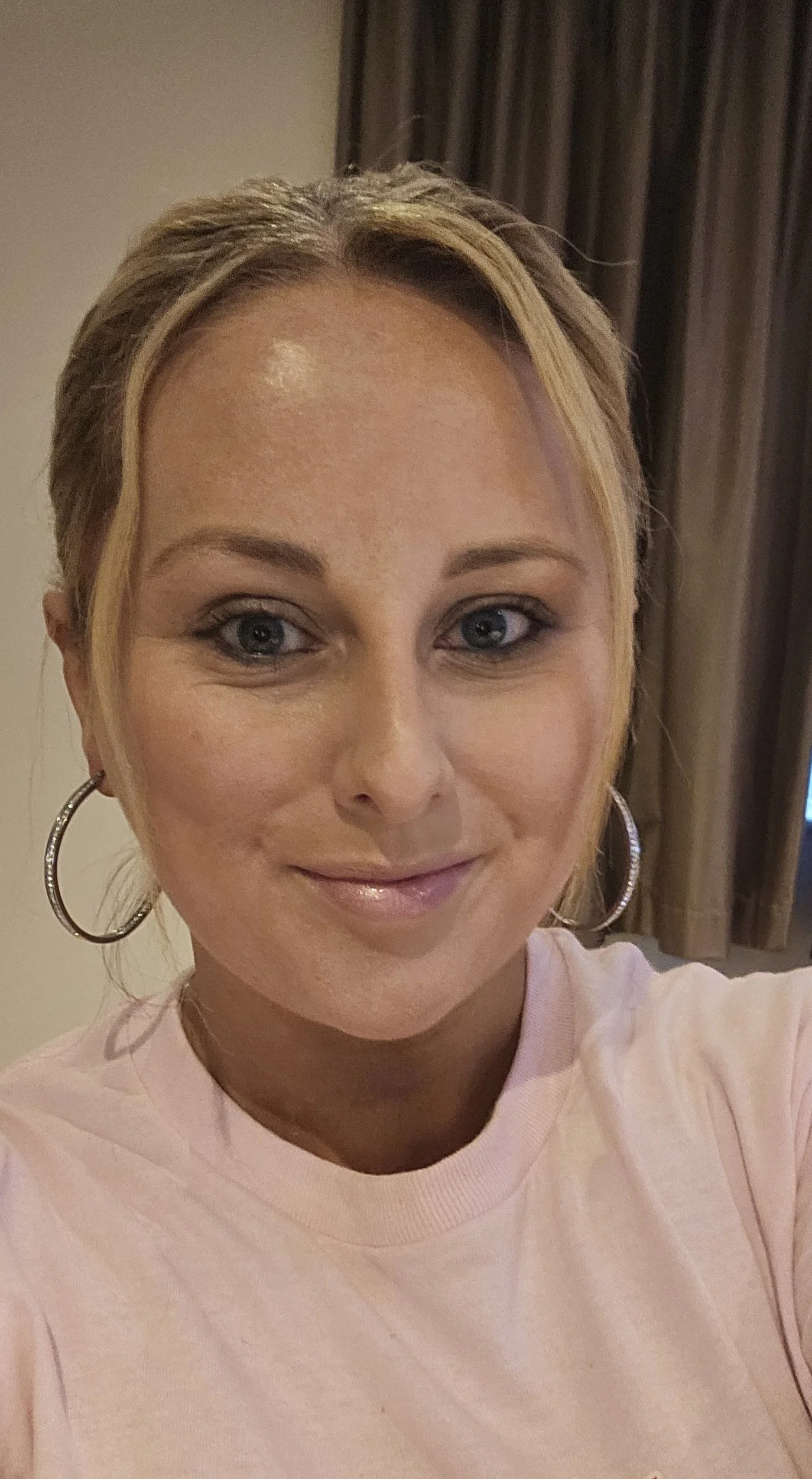 A woman with blonde hair, blue eyes, hoop earrings, and a light pink shirt, smiling in a room with beige walls and dark curtains.