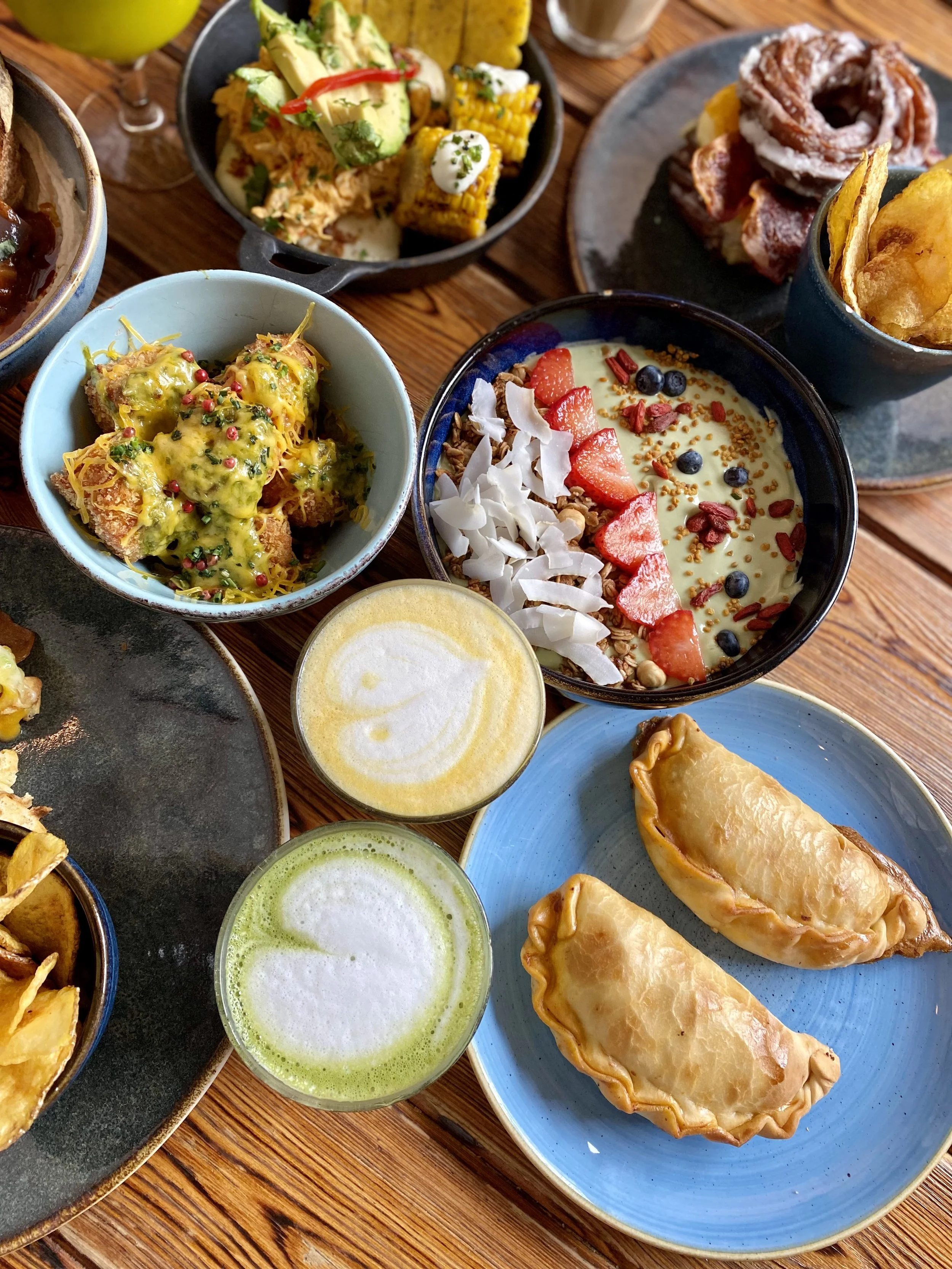 Comida variada que incluye empanadas, un tazón de fruta con yogur, una ensalada, diferentes dips y bebidas, todo presentado en tazones y platos sobre una mesa de madera.
