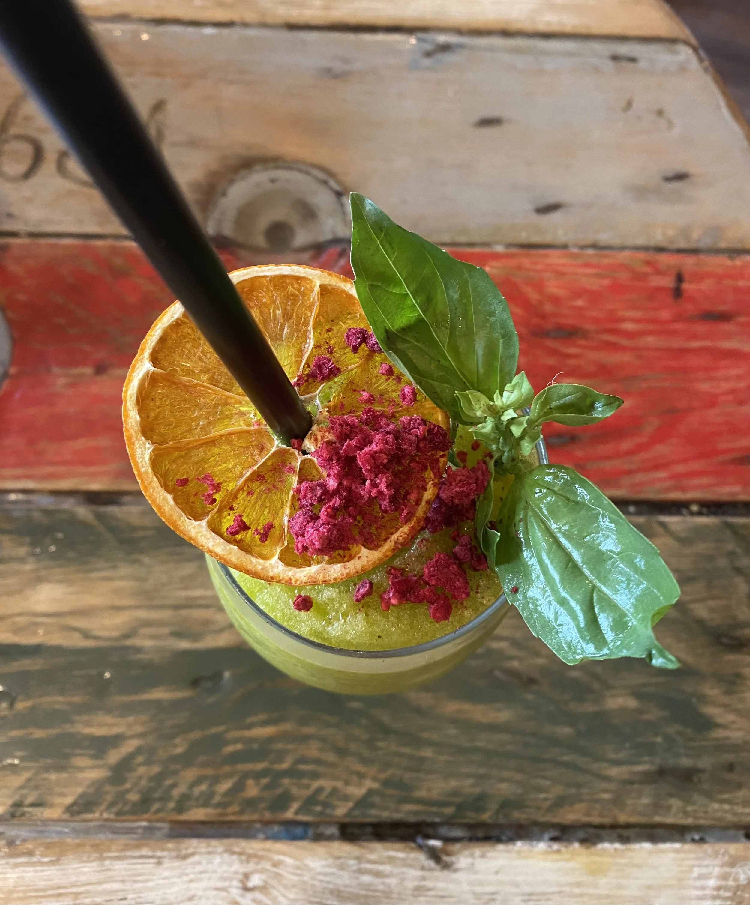 Bebida verde con rodaja de naranja, hojas de albahaca y polvo rosa en la parte superior, con un popote negro.