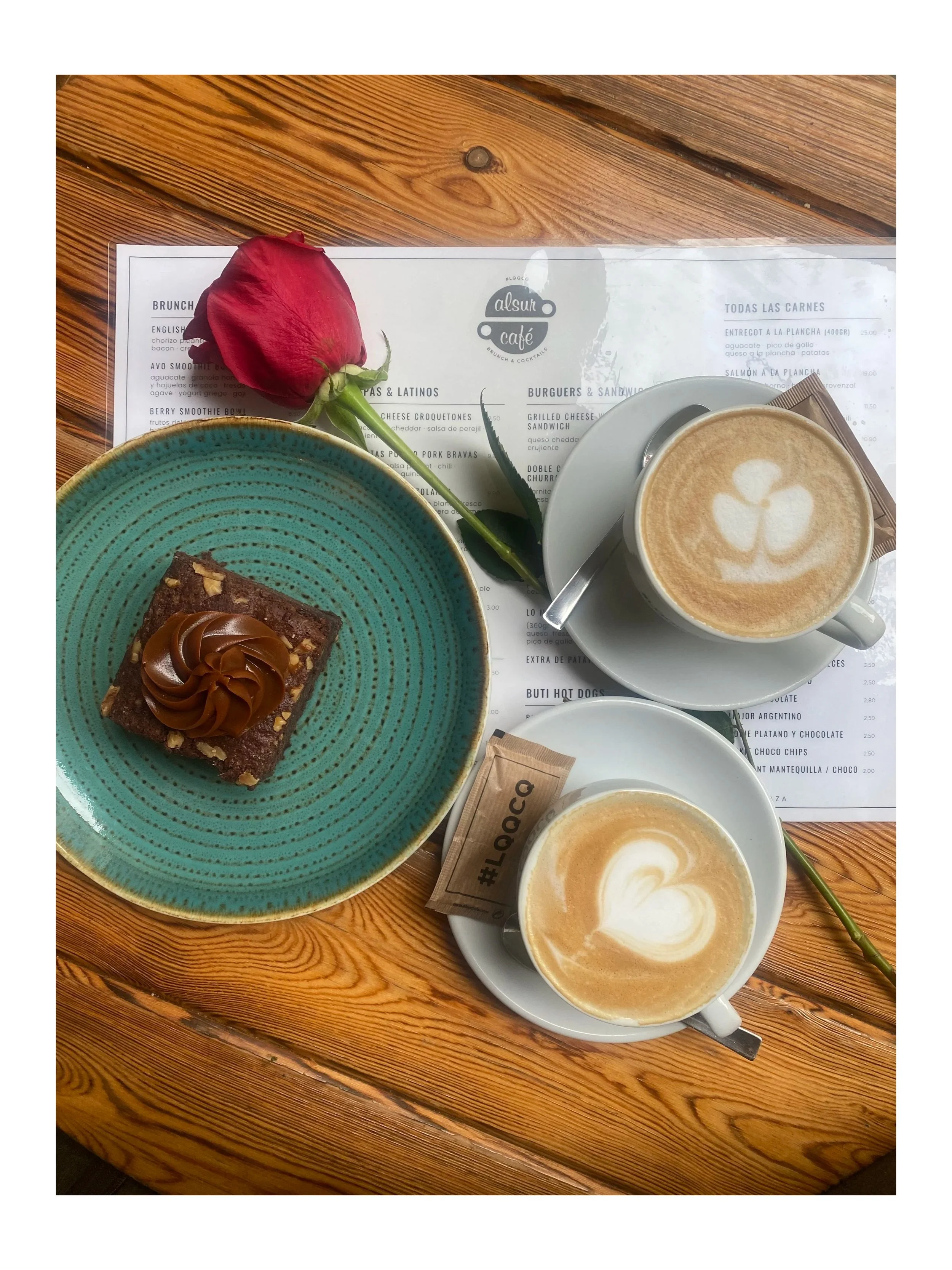 Una mesa de madera con dos tazas de café con arte en la espuma, un plato con un Brownie de chocolate decorado con dulce de Leche, una rosa roja y un menú en el fondo.