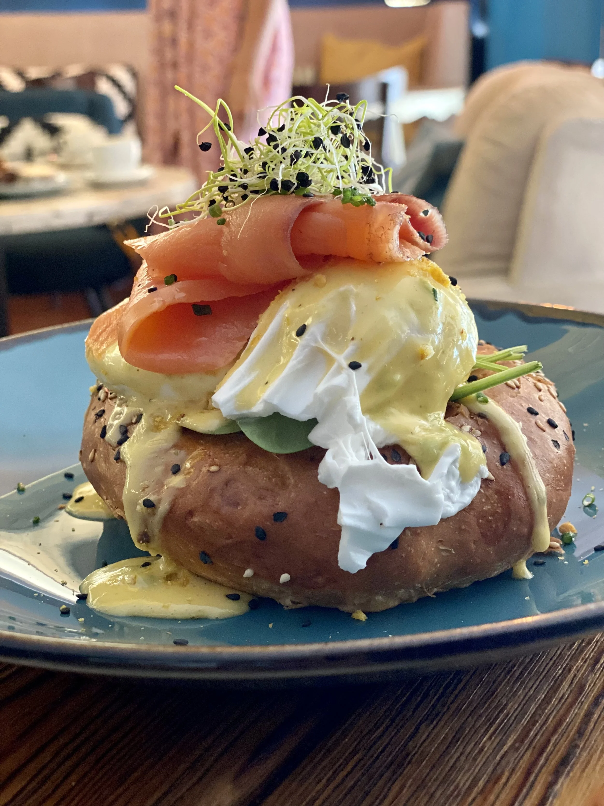 Huevos pochados sobre una papa, con jamón, microgreens y semillas negras condimentadas, en un plato azul.