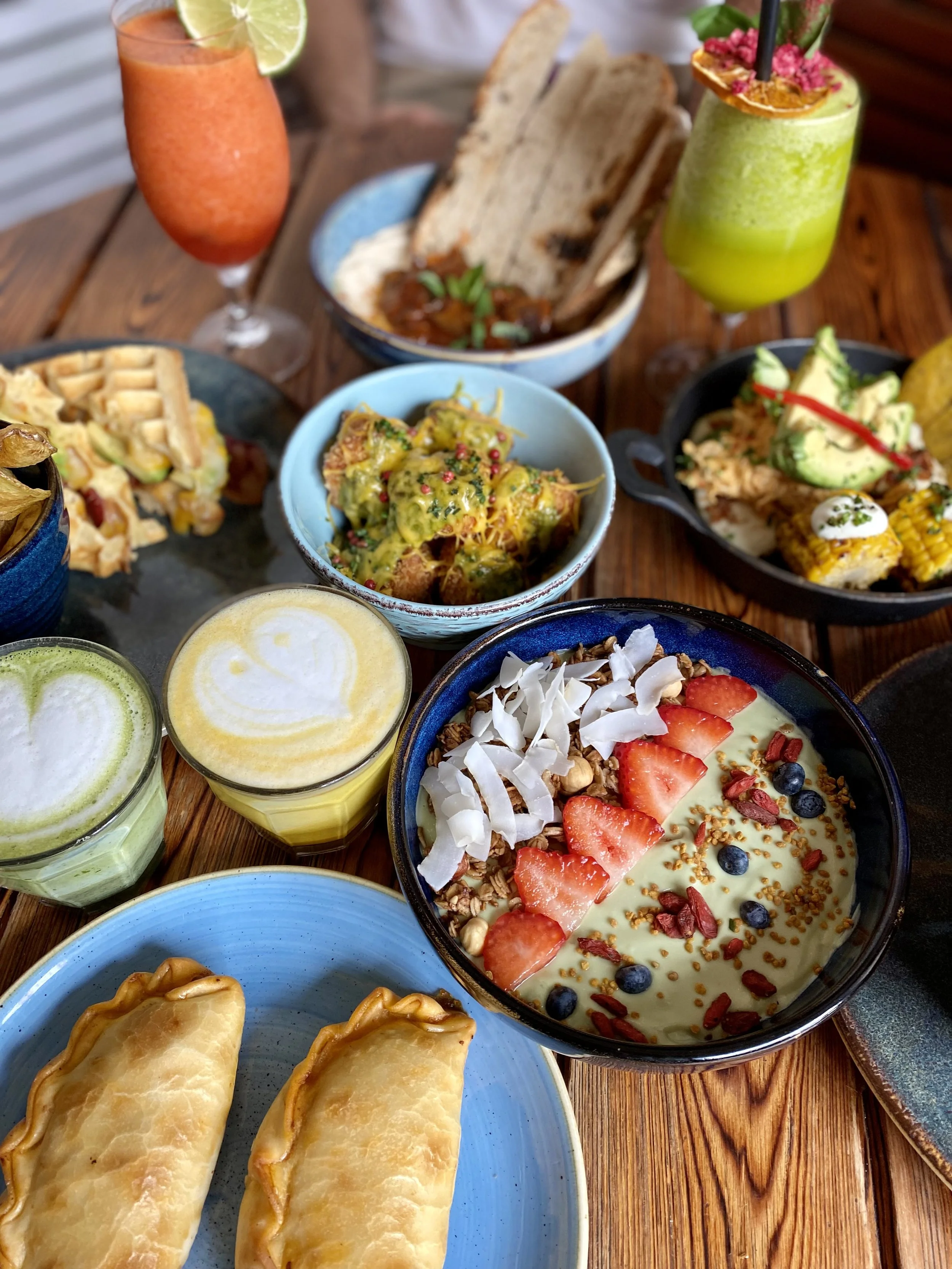 Una variedad de alimentos y bebidas en una mesa, incluyendo jugos, una sopa con pan, tacos, una ensalada de frutas y huevos, y cócteles coloridos.