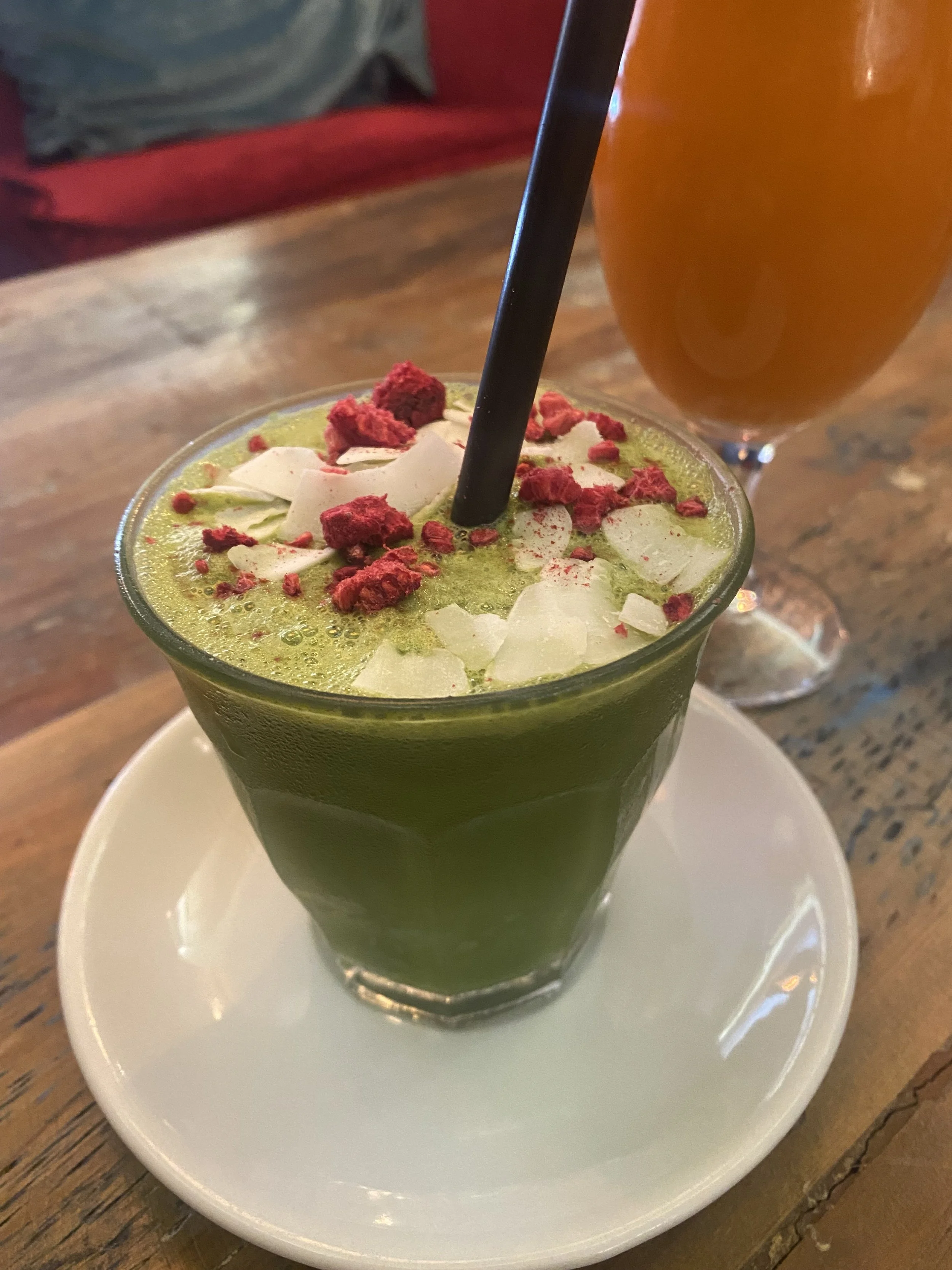 Vaso con jugo verde decorado con trozos de coco y polvo de fresa y un vaso con cerveza en el fondo en una mesa de madera.