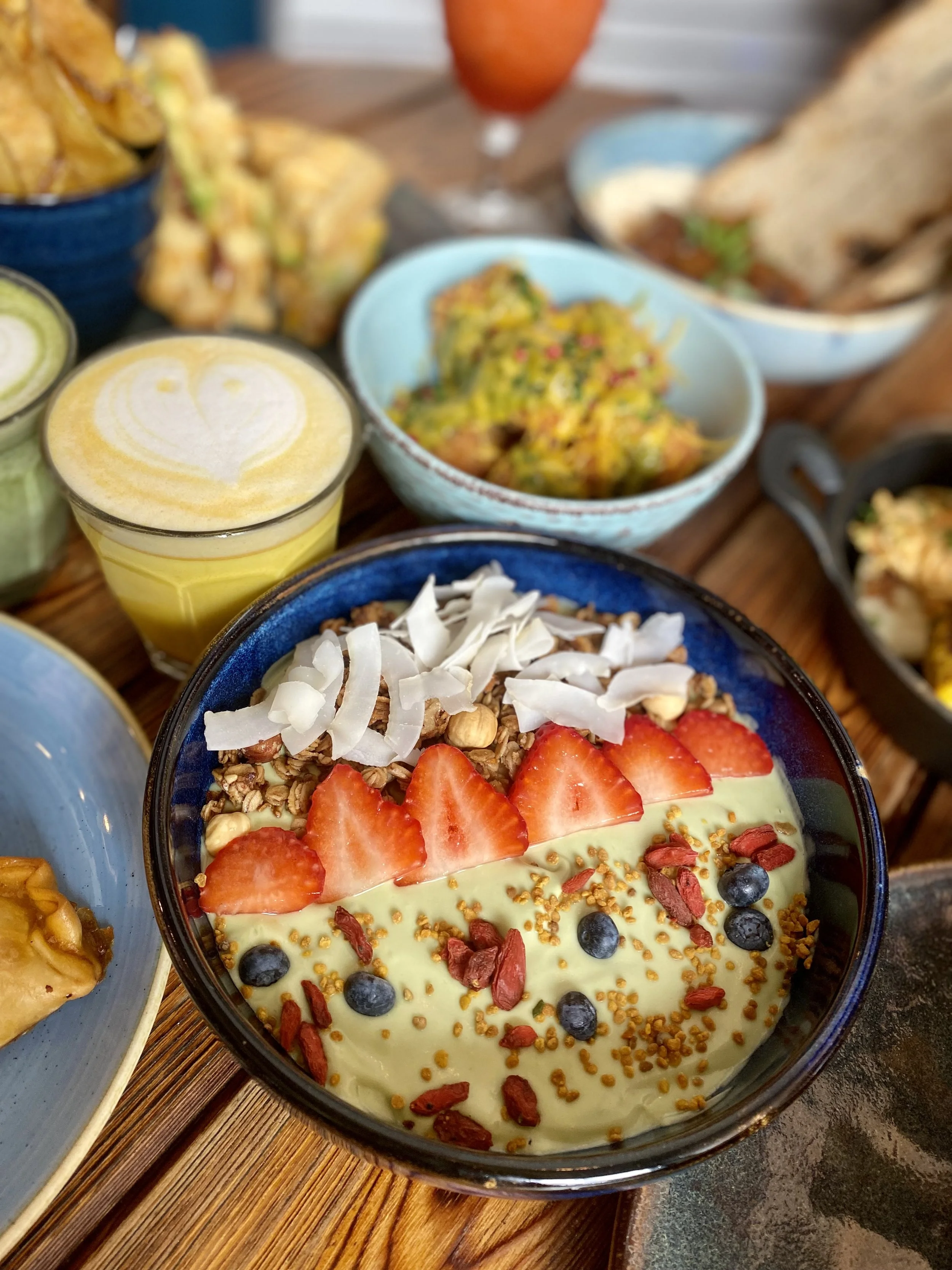 Ensalada de yogur con fresas, arándanos, nueces, coco rallado y semillas de chía en un bol negro, rodeada de otros platos y bebidas en una comida