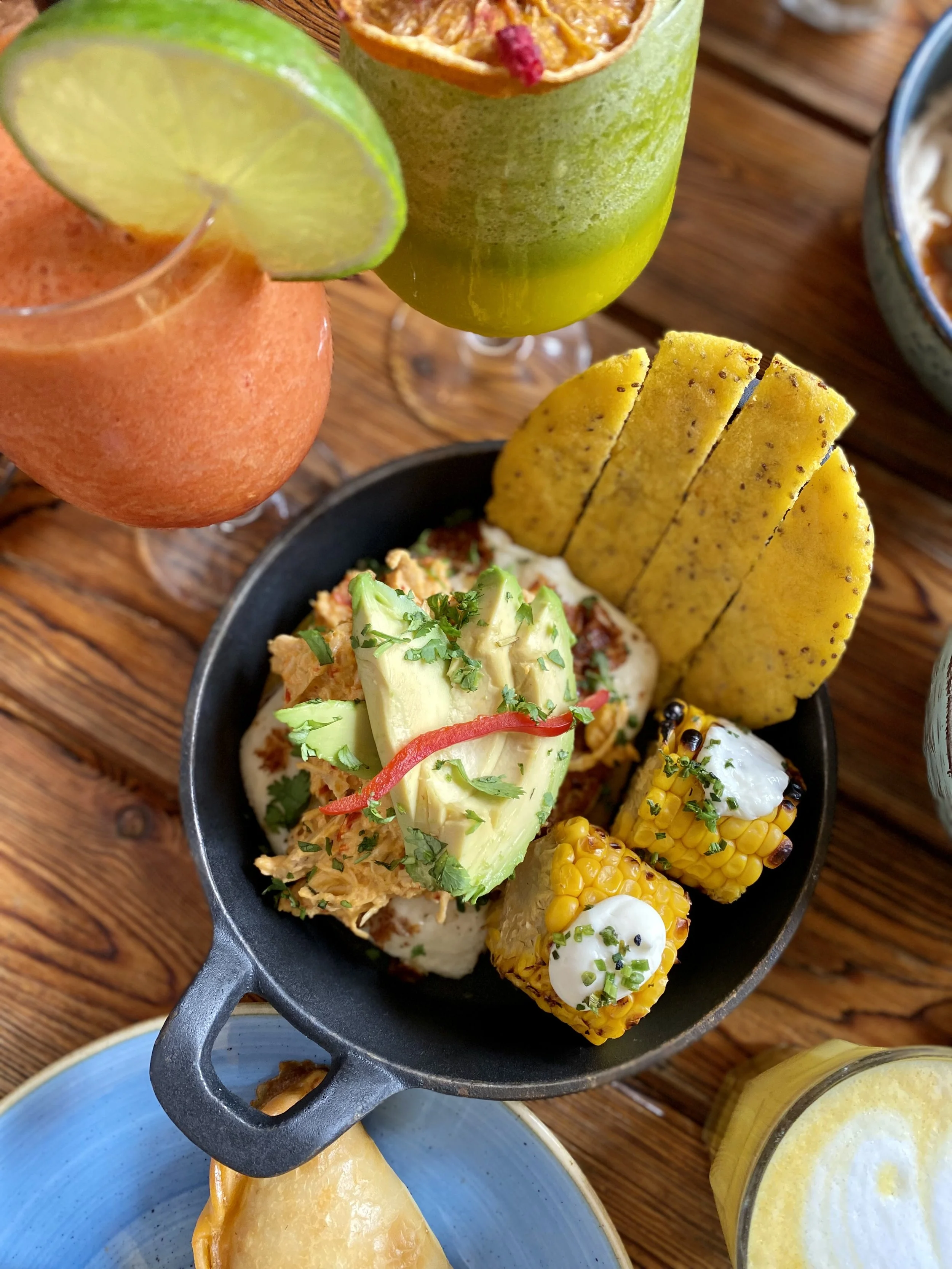 Plato con maíz, aguacate, cilantro, y totopos acompañado de bebidas con limón, aguacate, y otros ingredientes en el fondo.