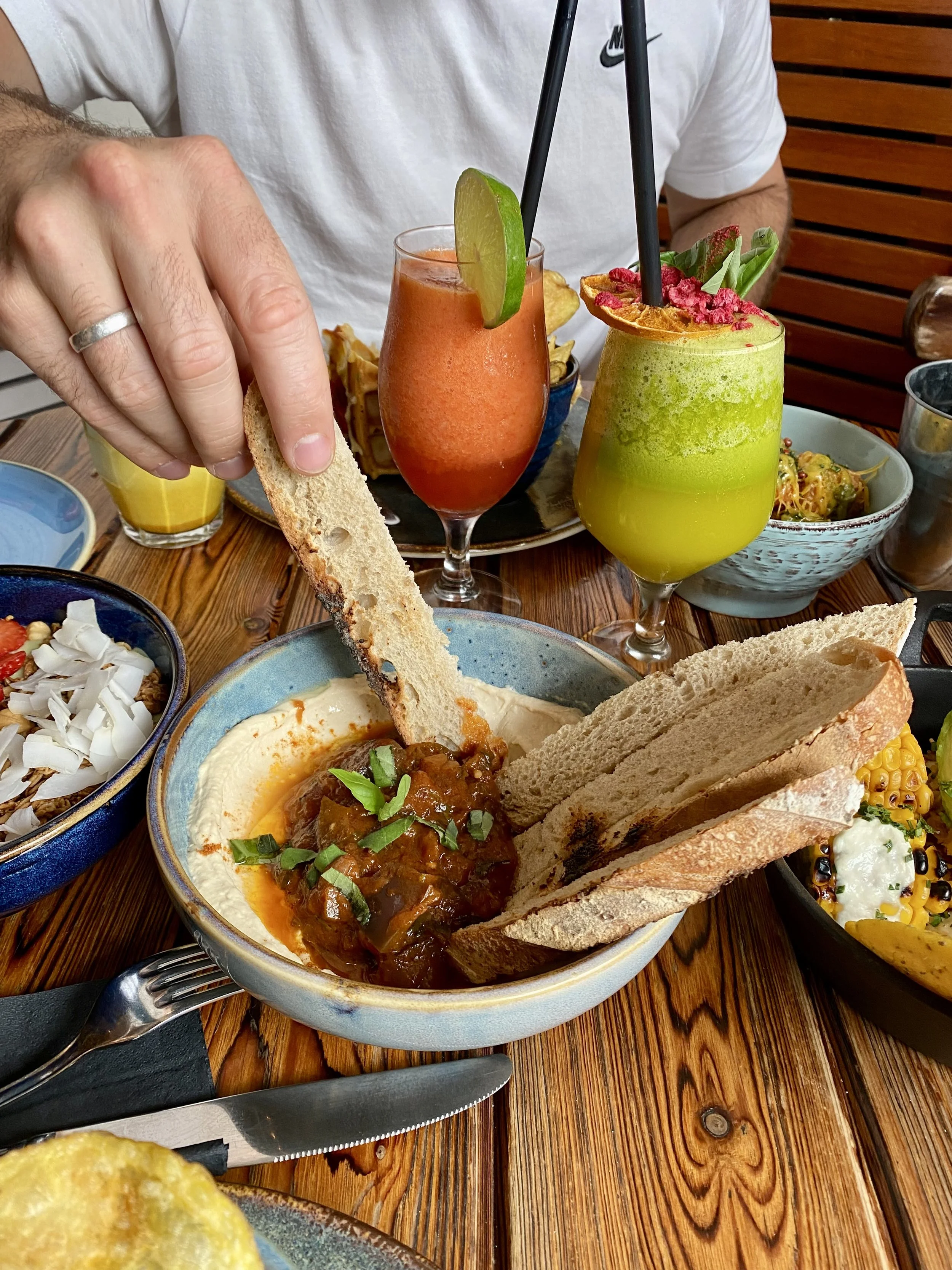 Plato con hummus, pan y salsa de carne en un plato azul. En el fondo, bebidas coloridas y personas en una mesa de madera.