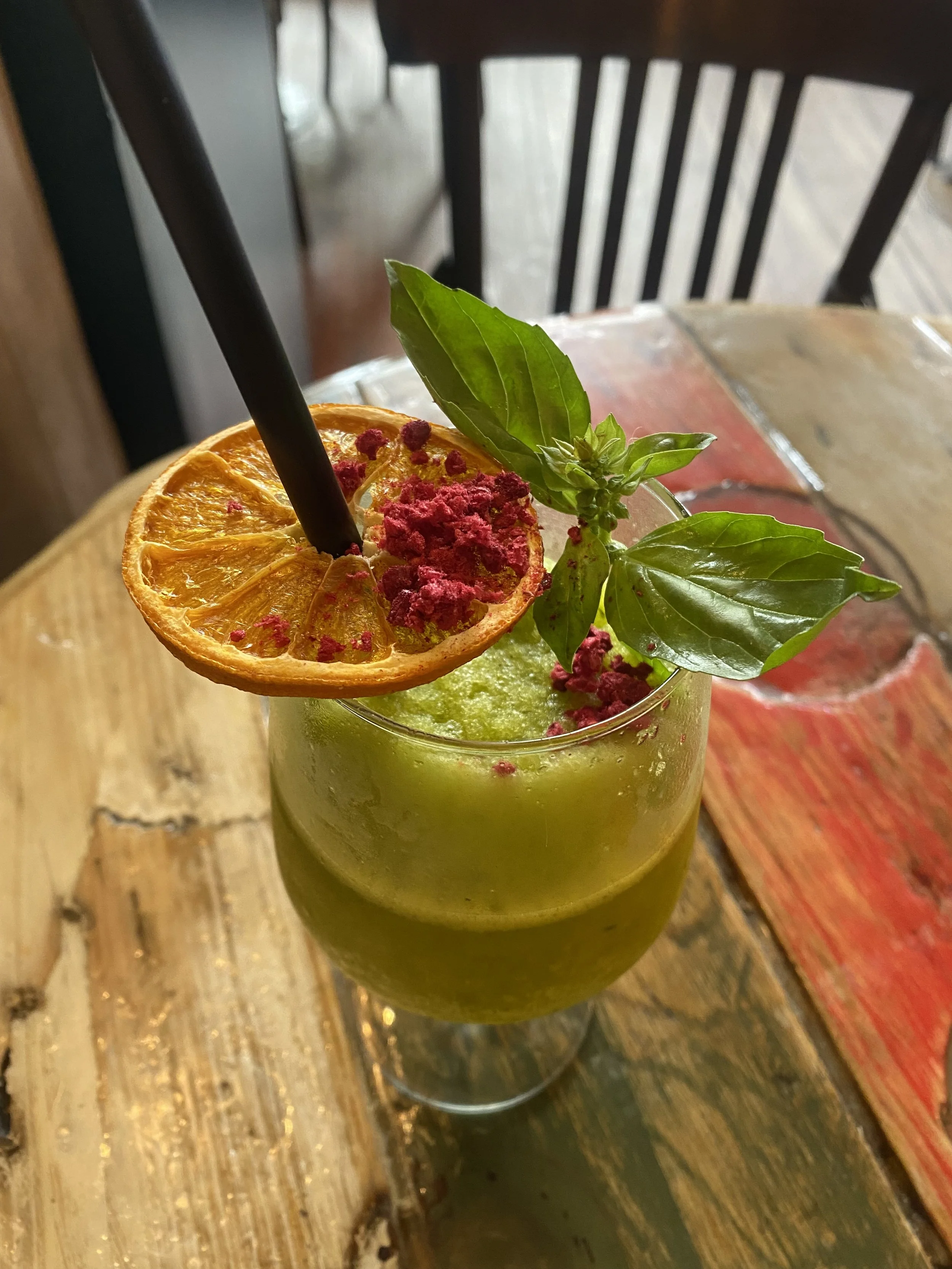 Cóctel verde decorado con una rodaja de naranja, hojas de albahaca y polvo rosa, en un vaso alto en una mesa de madera.