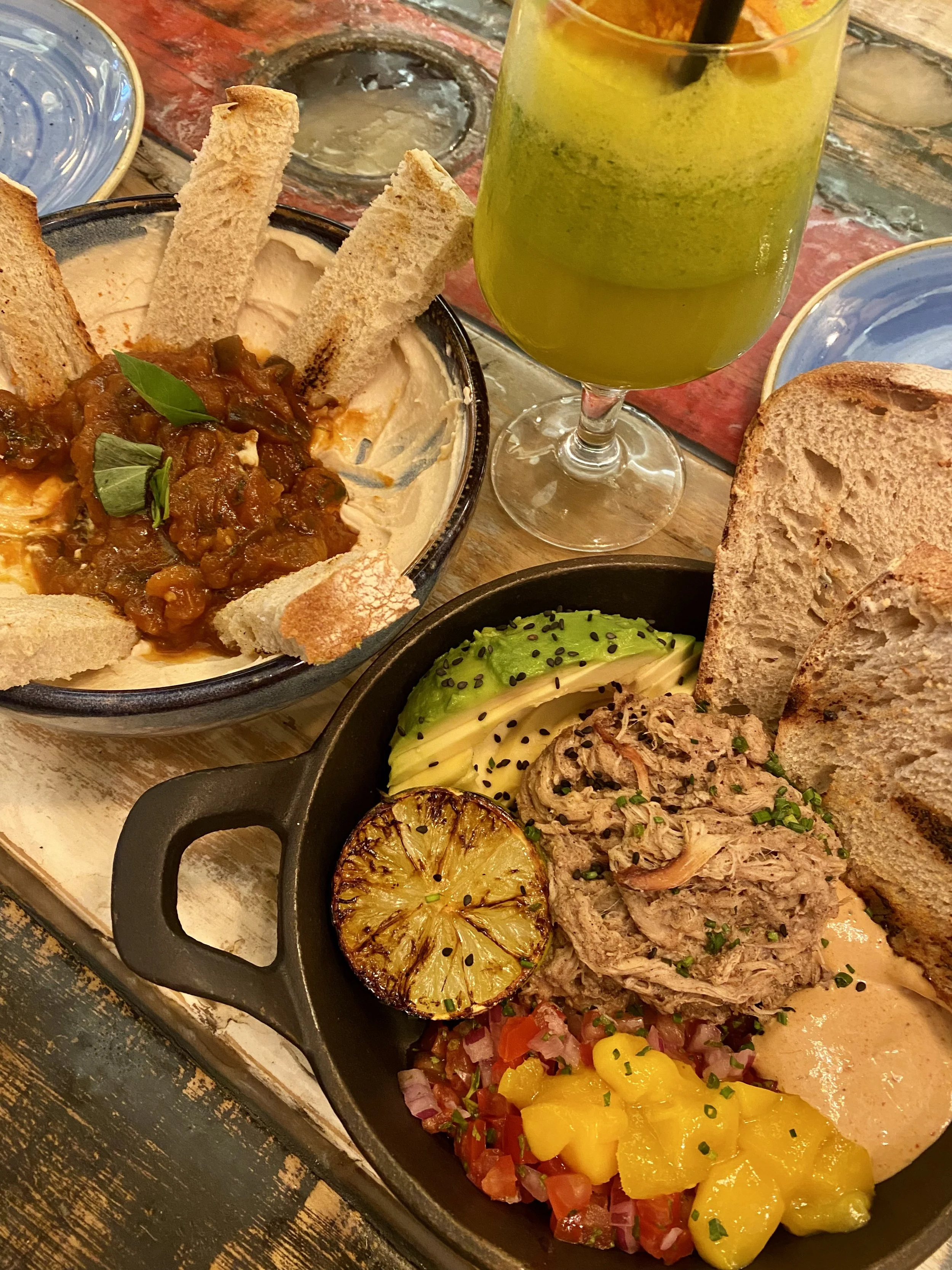 Plato de comida con hummus cubierto de salsa de tomate con pan frito, guacamole con aguacate, limón asado, verduras picadas y pan en un plato."