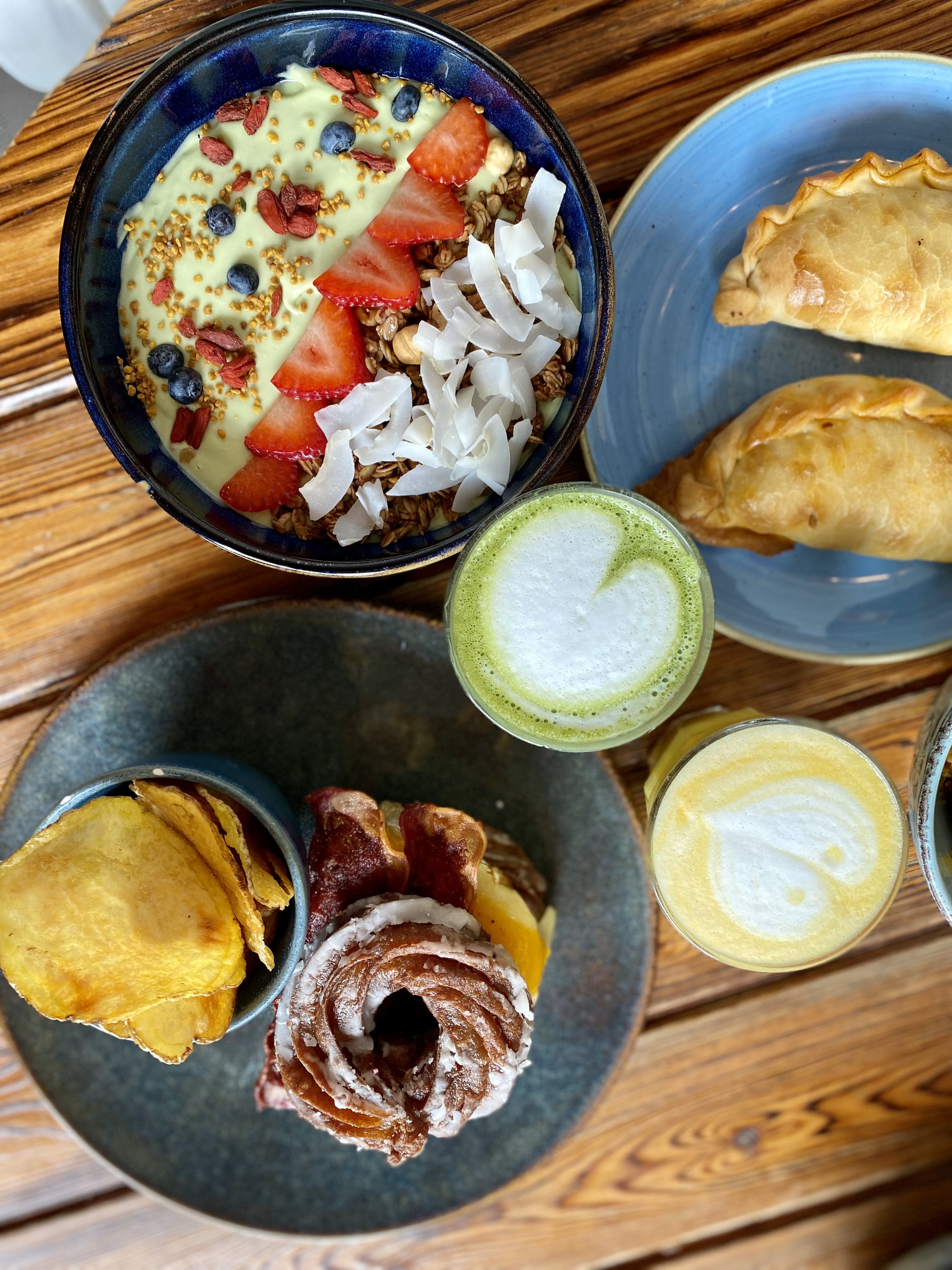 Desayuno con yogur con frutas, granola, gofres, chips de plátano y bebidas verdes y amarillas en vasos en una mesa de madera.