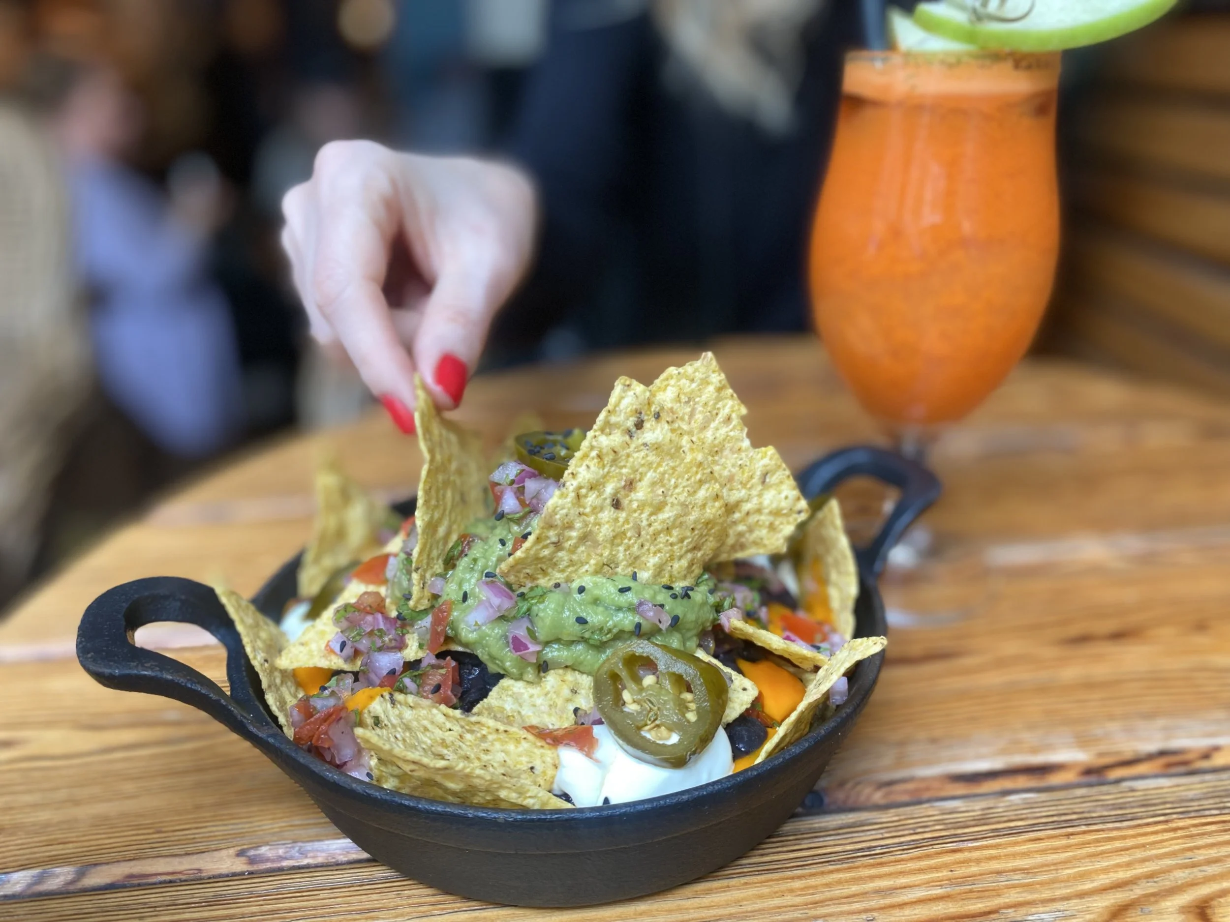 Tazón de nachos con guacamole, jalapeños, pico de gallo y crema, en una mesa de madera, con una bebida en un vaso largo al fondo.