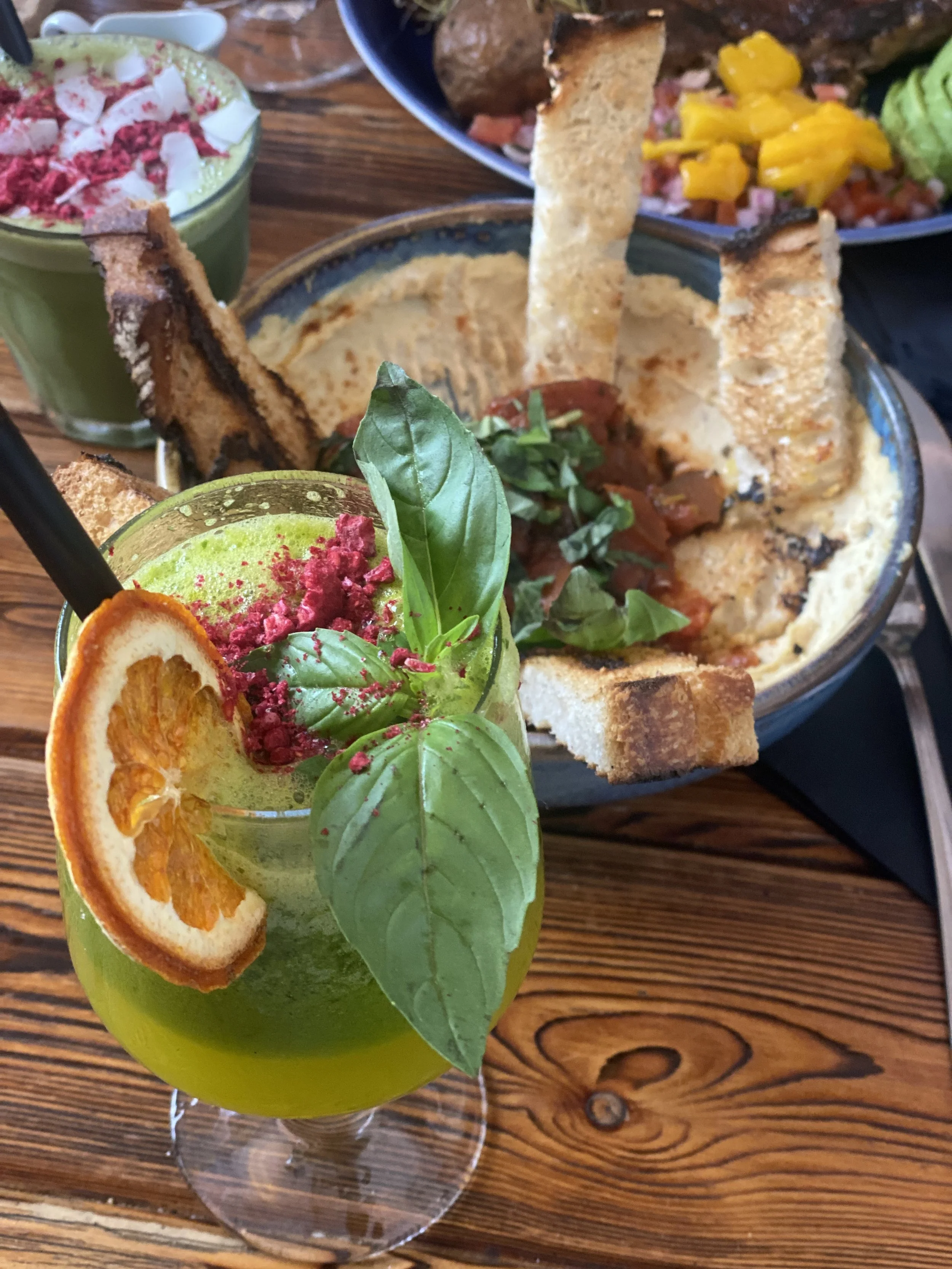 Cóctel verde con hojas de albahaca, rodaja de naranja, y polvo rosa, junto a comida con pan tostado, salsa y ensalada en platos. Fondo de mesa de madera.