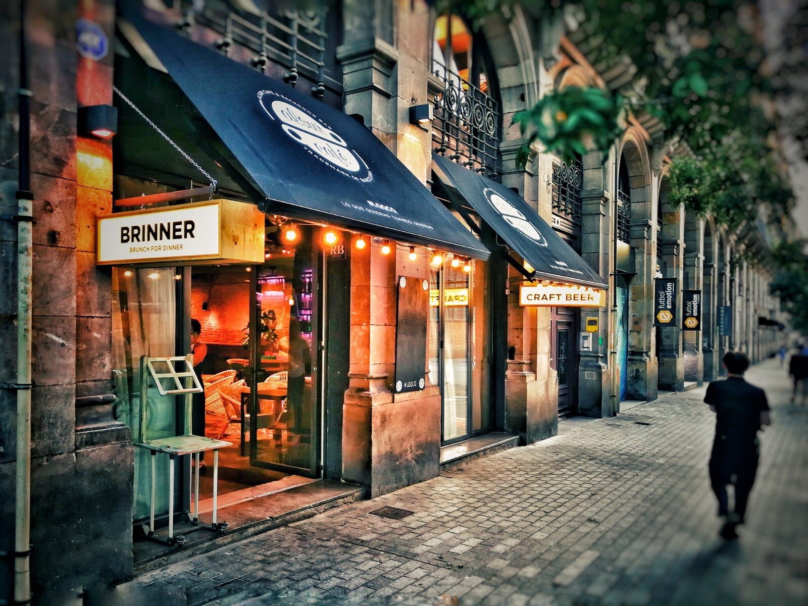 Vista de un restaurante o bar en la calle con cartel que dice "Brinner" y un letrero de "Craft Beer". La fachada está iluminada con luces cálidas, con varios carteles y ventanas que muestran la decoración interior. En la acera hay una persona que cam