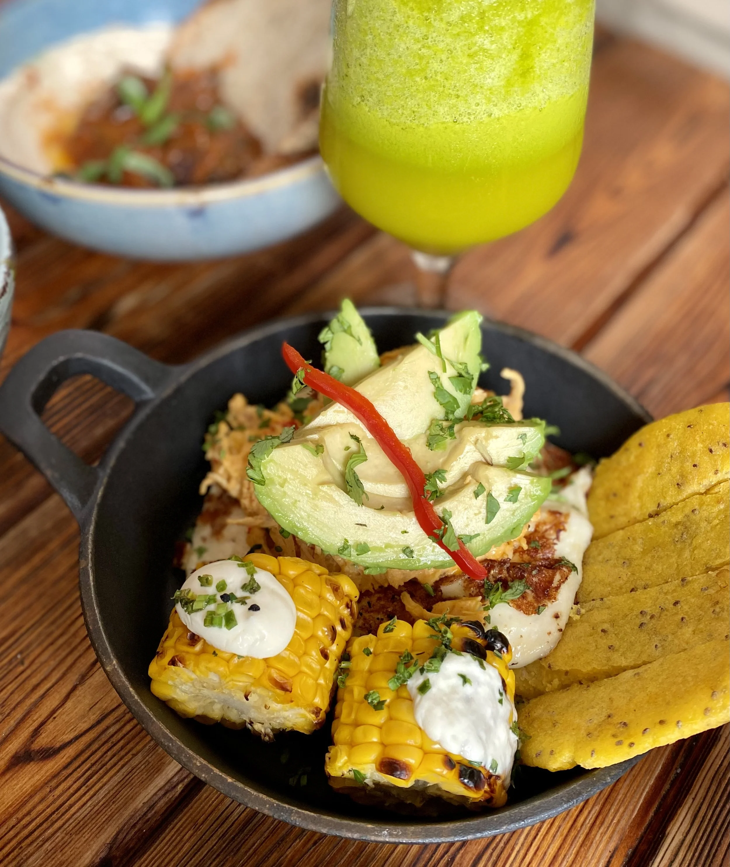 Plato de elote con mayonesa, cilantro y cebolla, acompañado de totopos, guacamole y salsa, con una bebida verde al fondo.