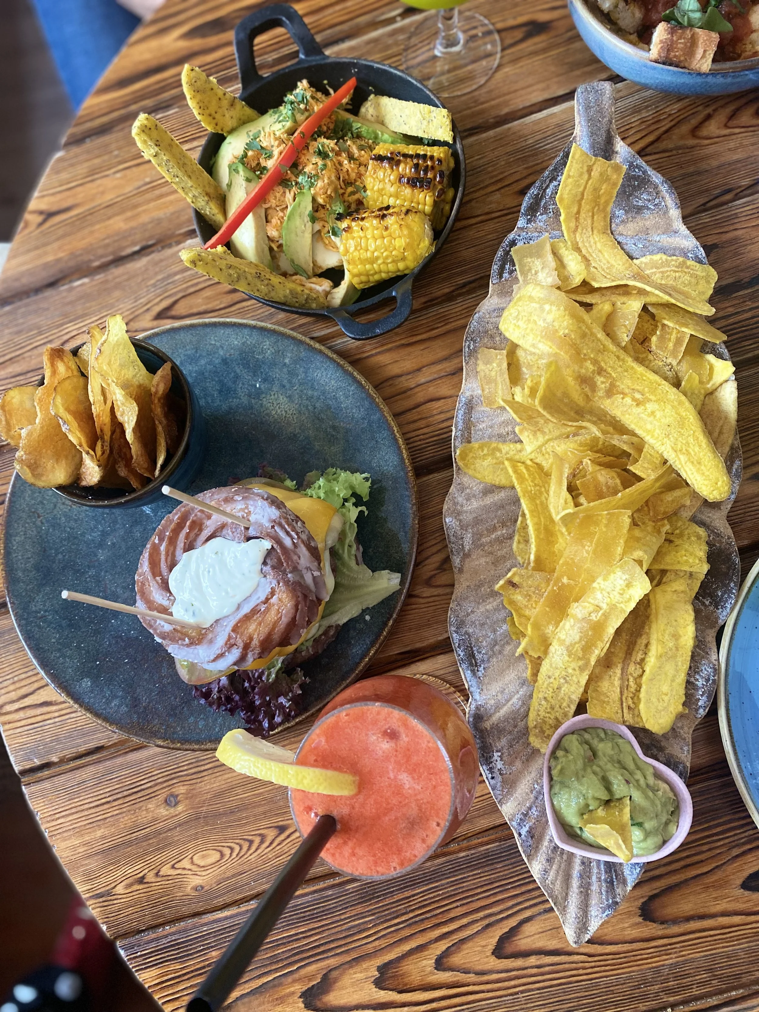 Variedad de alimentos mexicanos en la mesa, incluyendo totopos, guacamole, una hamburguesa, una bebida con rodaja de limón, y una ensalada con maíz a la parrilla.