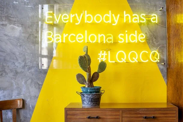 Pared de fondo con letrero de neón en inglés que dice 'Everybody has a Barcelona side #LQGCO', decoración con cactus en una maceta sobre una mesa de madera, y el fondo de la pared es gris con una sección amarilla en forma de 'V'.