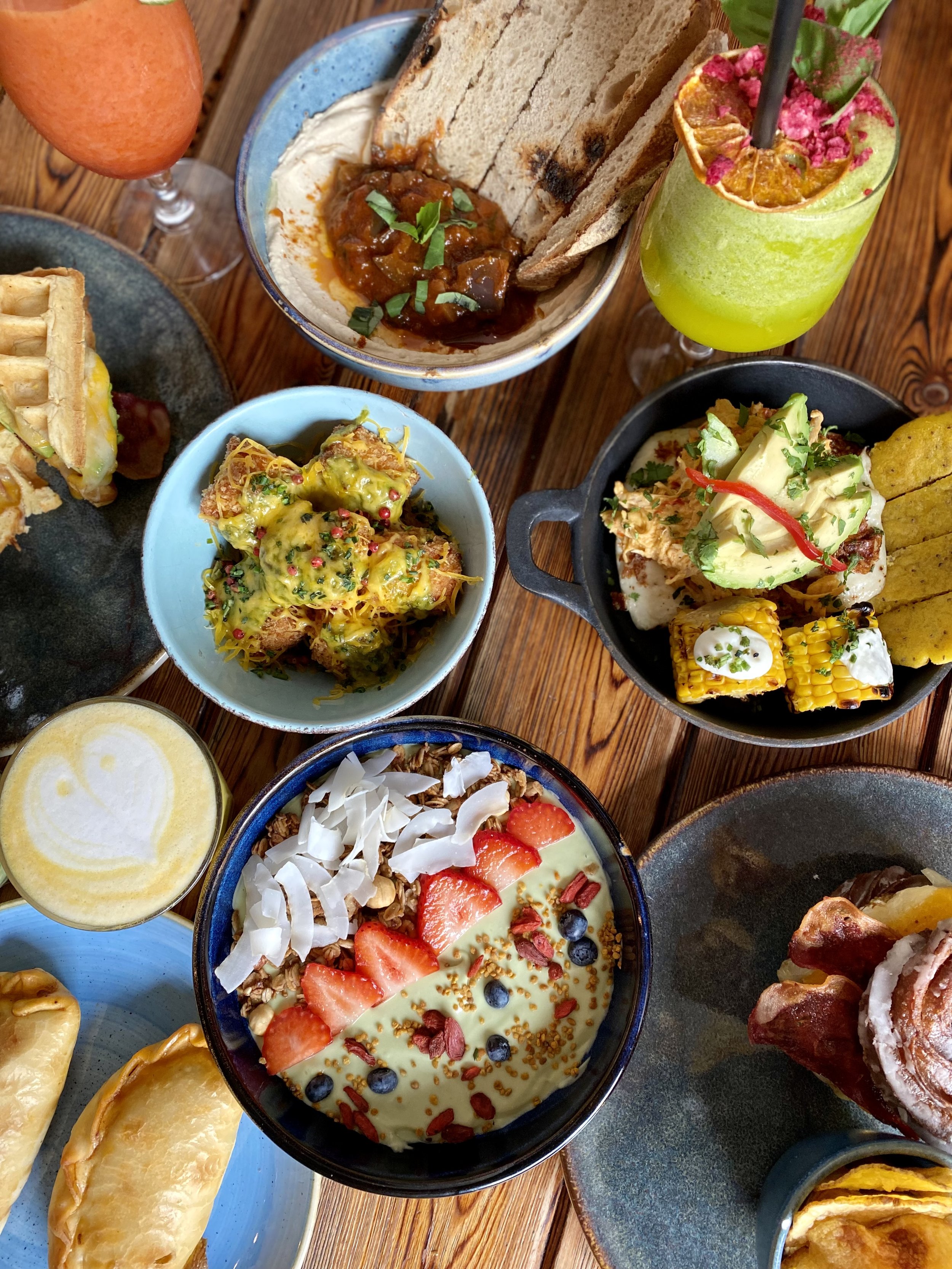 Variedad de platos y bebidas de comida y postres, incluyendo pan, ensaladas, un smoothie de color verde, y un bowl de yogurt con frutas y semillas, en una mesa de madera.