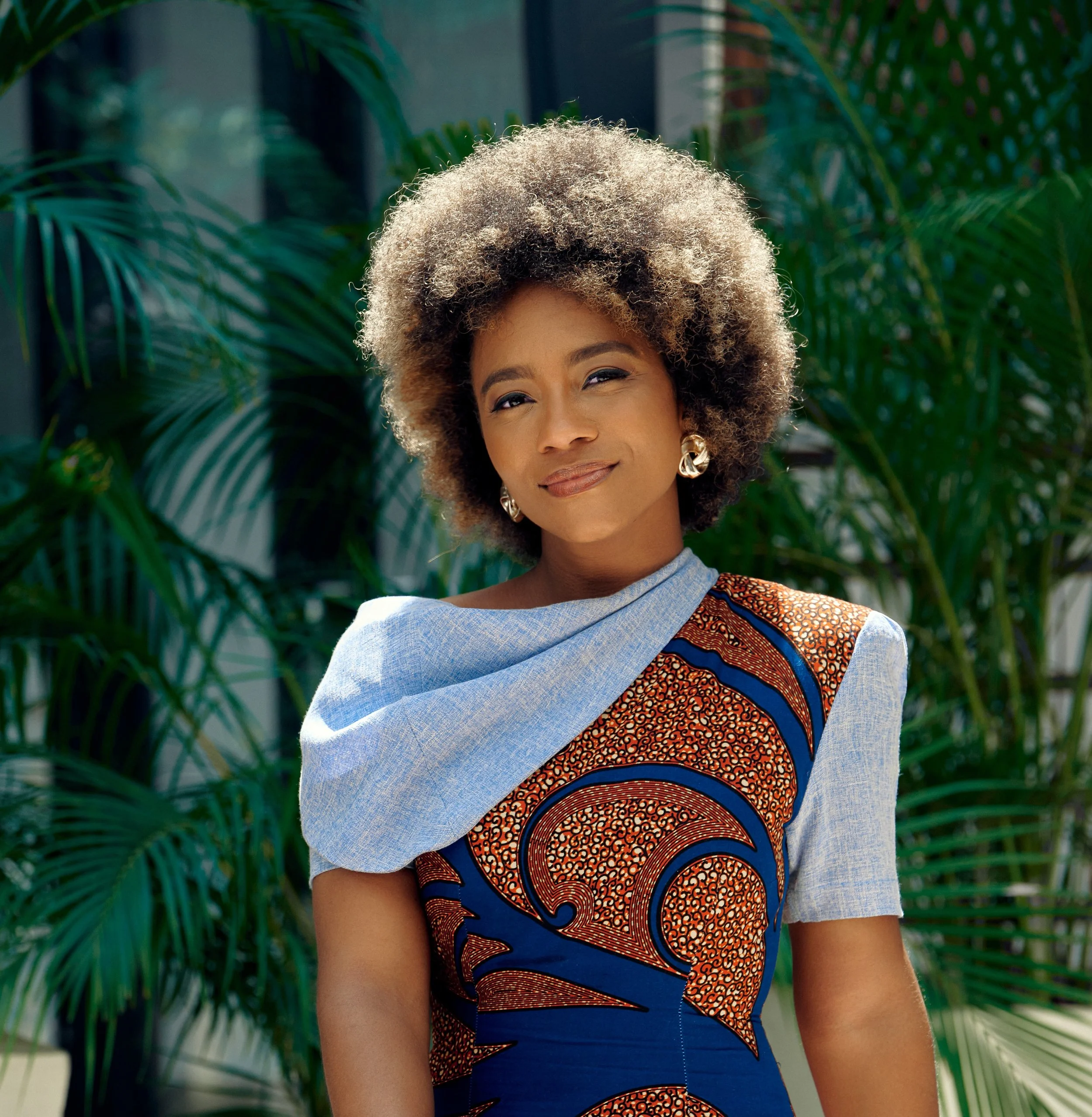 A woman with natural, curly hair wearing a colorful African print dress with a light blue scarf draped over her shoulder, standing outdoors with lush green plants in the background.
