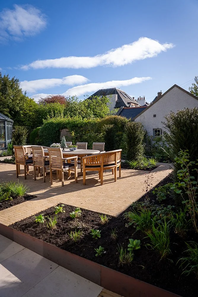 A backyard patio with a wooden dining table and chairs, surrounded by a garden with plants and shrubs, under a blue sky with white clouds.