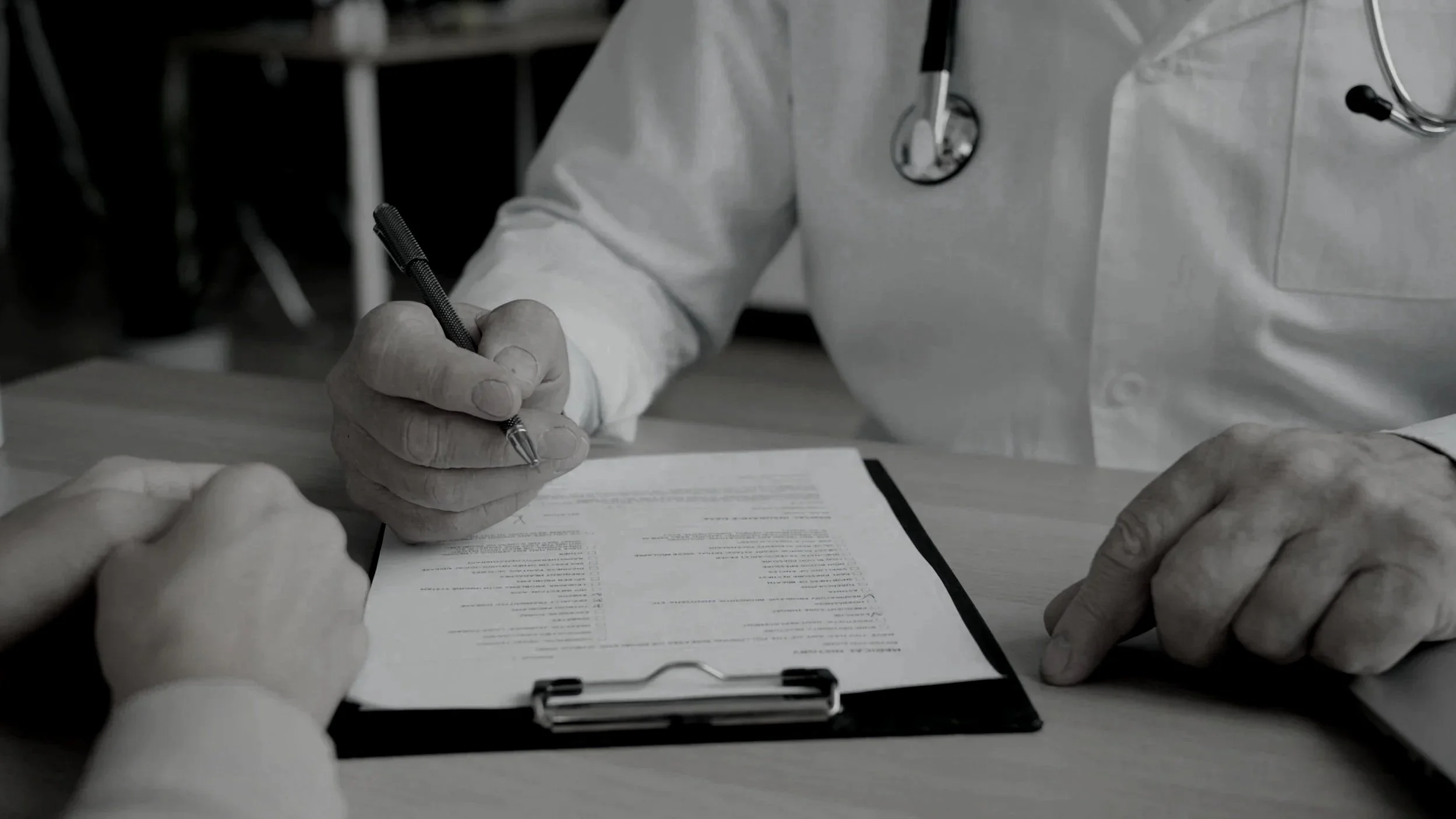 A healthcare professional in a white coat and stethoscope signs a document on a table with a patient's hand visible in the foreground.