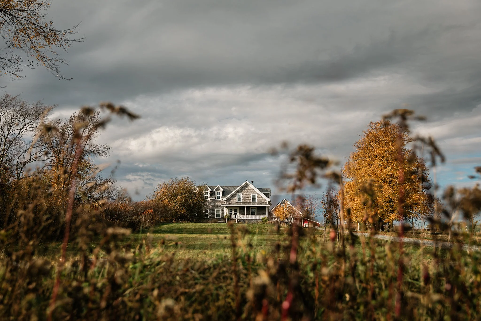 A large gray house with a green roof set on a hill, surrounded by trees with autumn foliage under a cloudy sky.