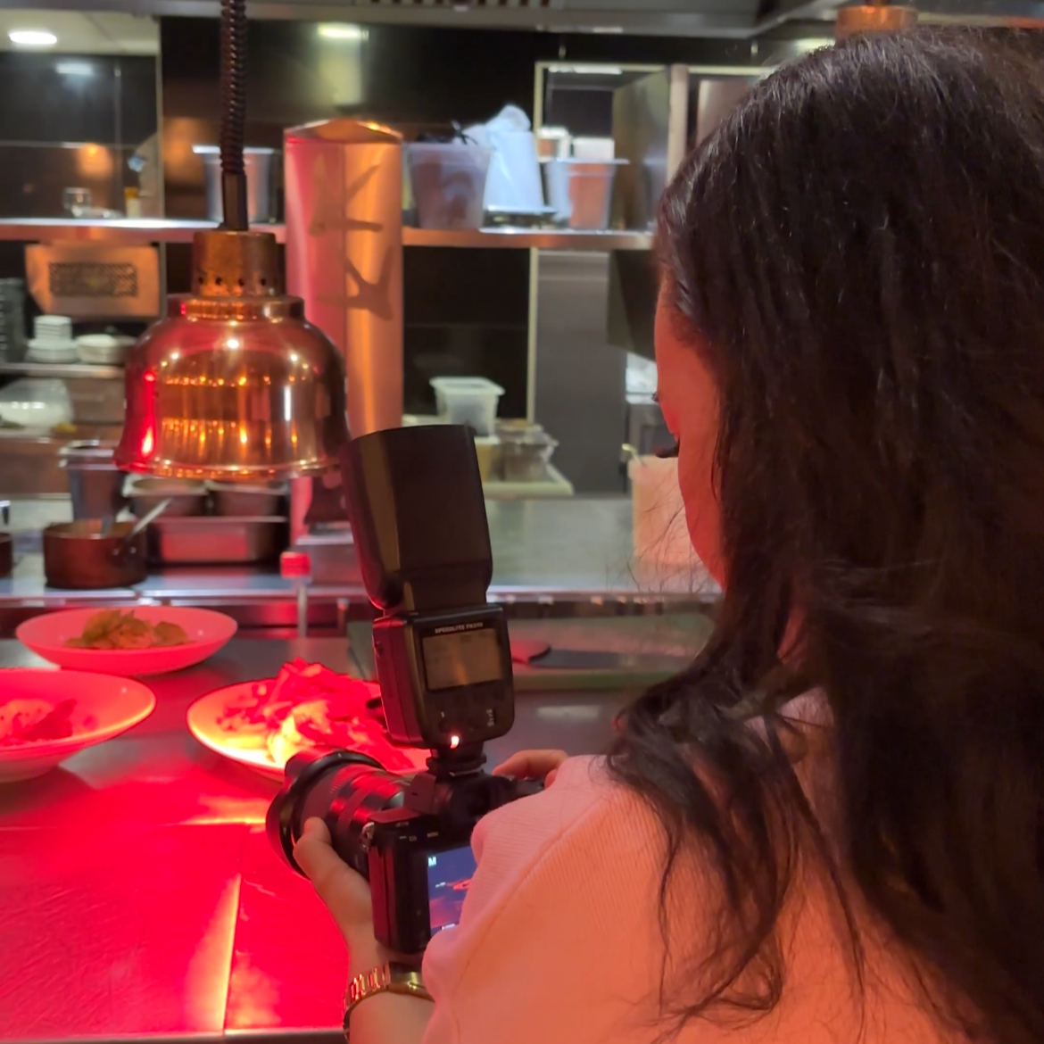 Une femme prenant une photo dans une cuisine de restaurant, avec des plats et une lampe en métal en arrière-plan.