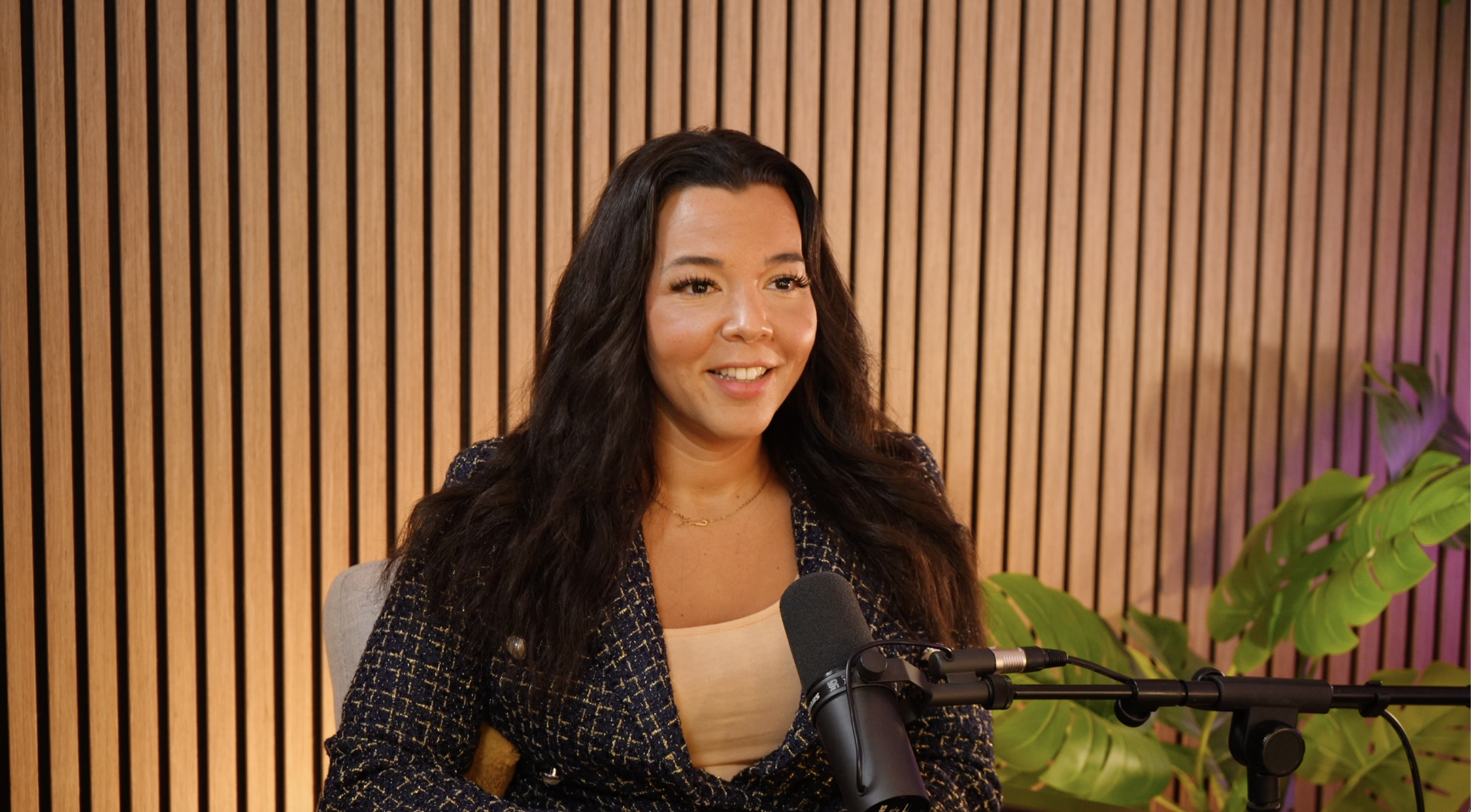 Femme souriante parlant dans un studio avec un microphone, fond en bois avec des plantes vertes.